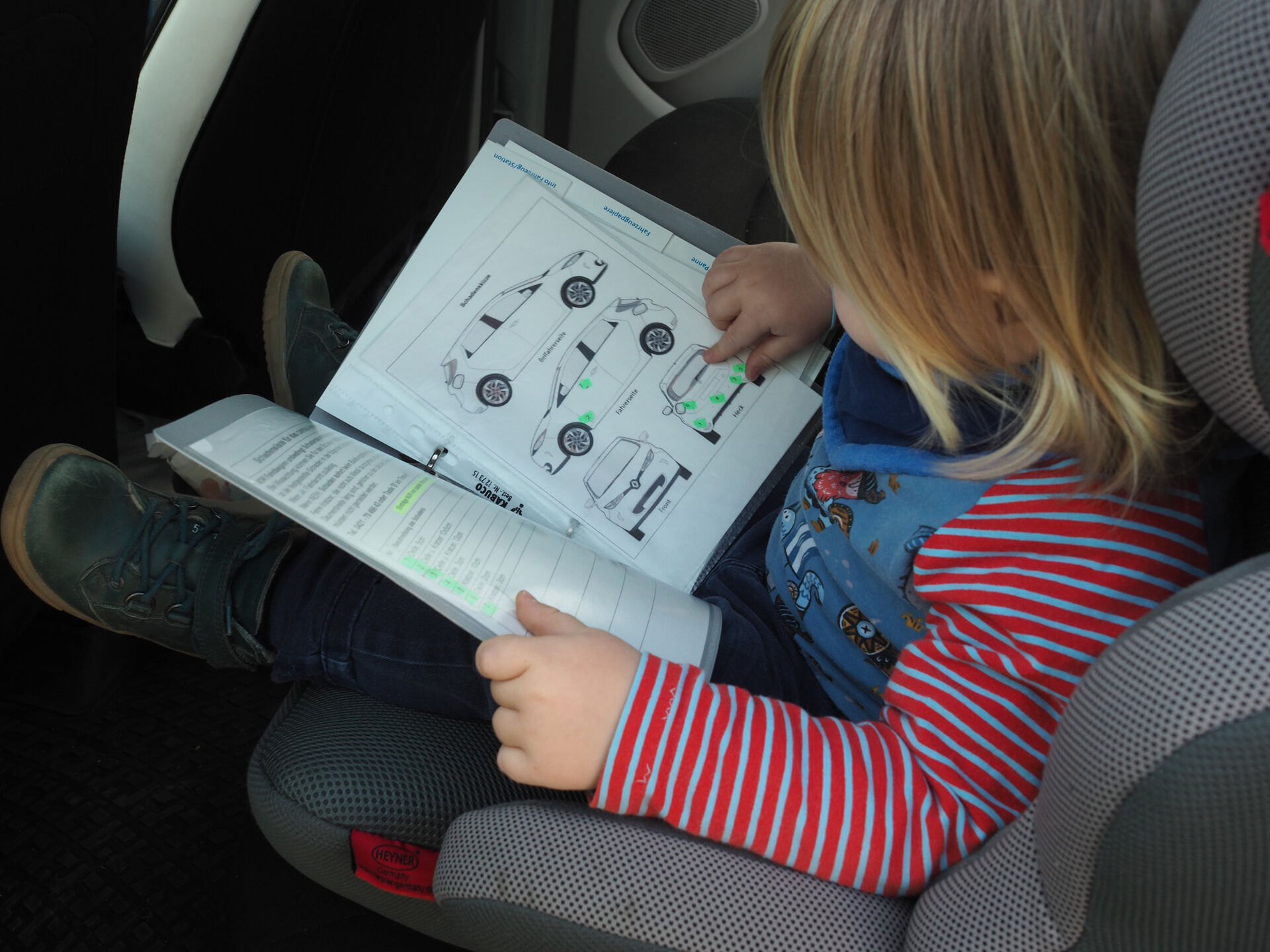 Ein Kind im Kindersitz im Auto, man schaut auf den Schoß. Darin liegt das Bordbuch, welches das Kind interessiert anschaut.