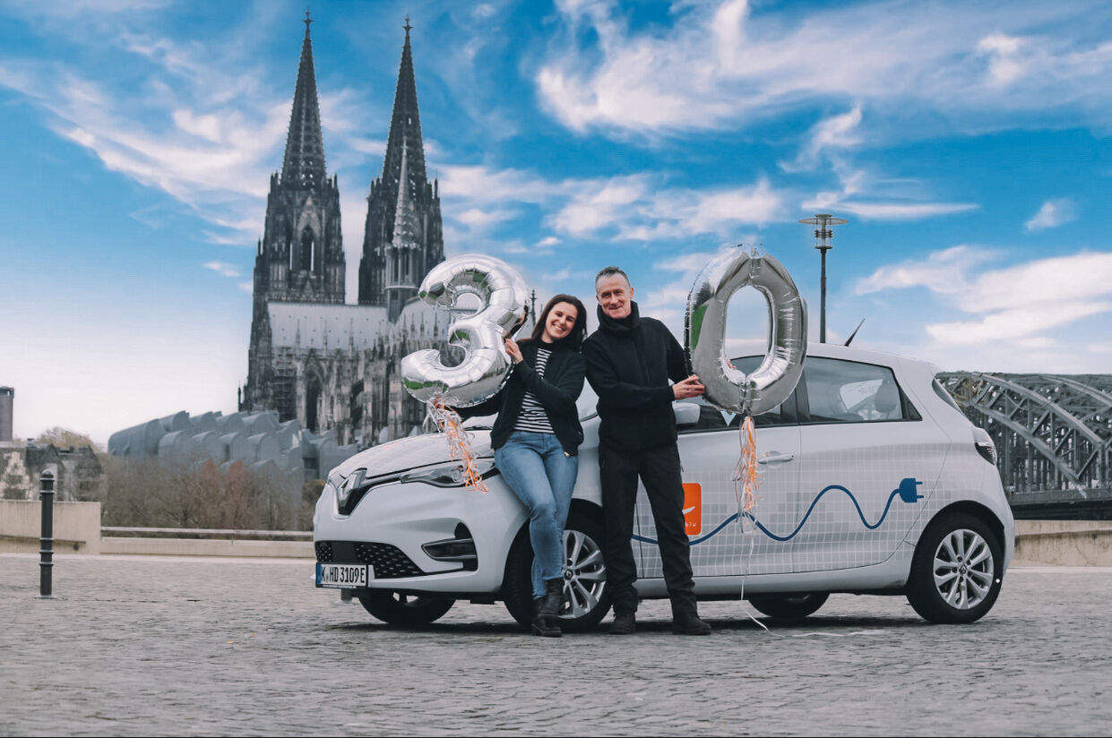 Eine Frau und ein mann stehen vor einem cambio-Auto. Sie halten Luftballons in die Höhe. Im Hintergrund der Kölner Dom.