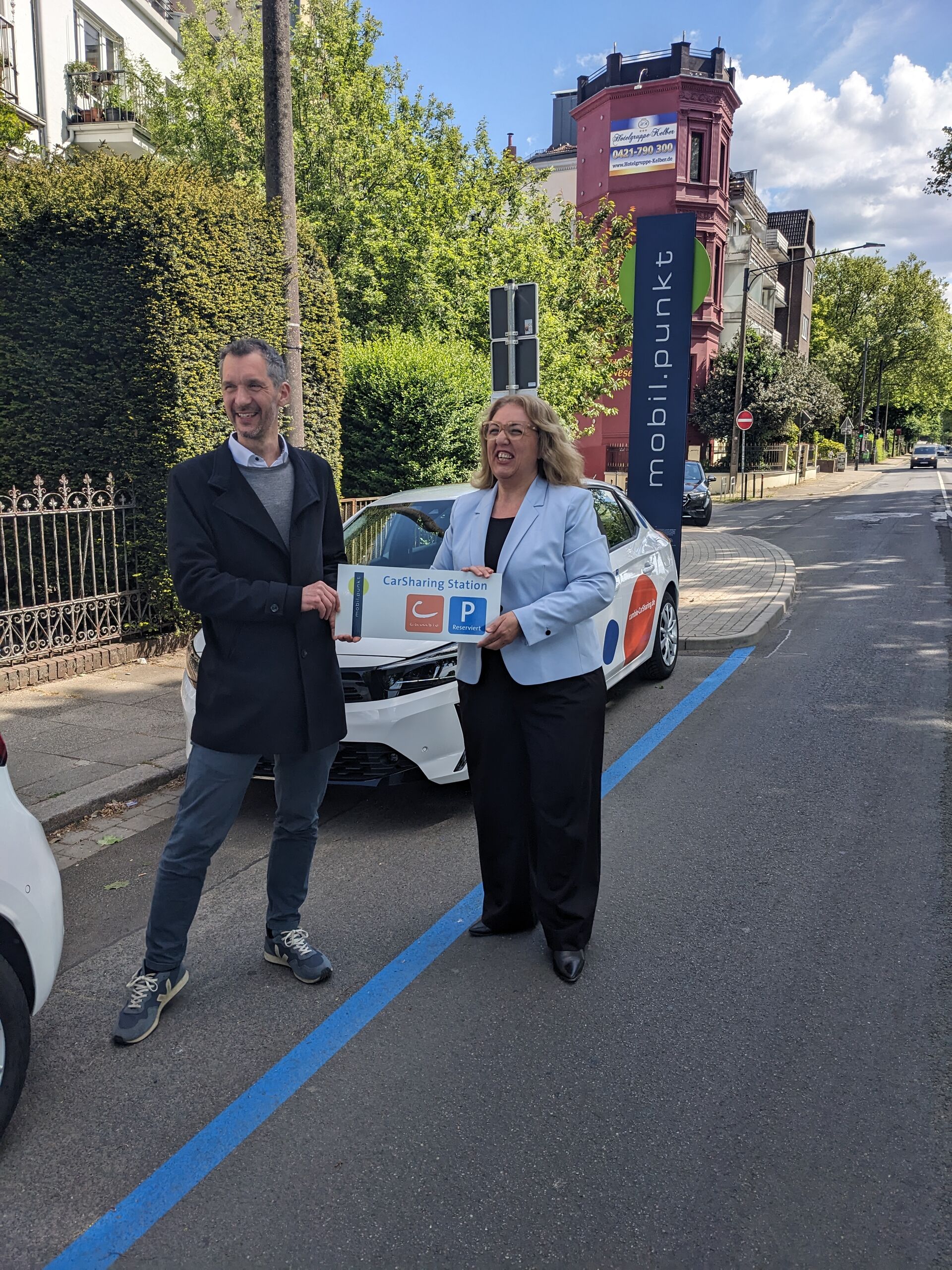 Lasse Schulz von cambio und Senatorin Özlem Ünsal halten ein Schild hoch, auf dem "CarSharing-Station" steht. Im Hintergrund sind die CarSharing-Station, Häuser und Bäume zu sehen.