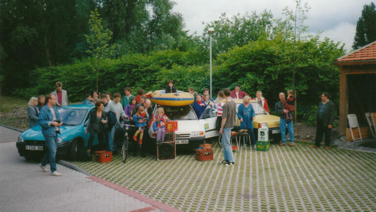 Ein Parkplatz mit zwei geparkten Autos und vielen Menschen drumherum.