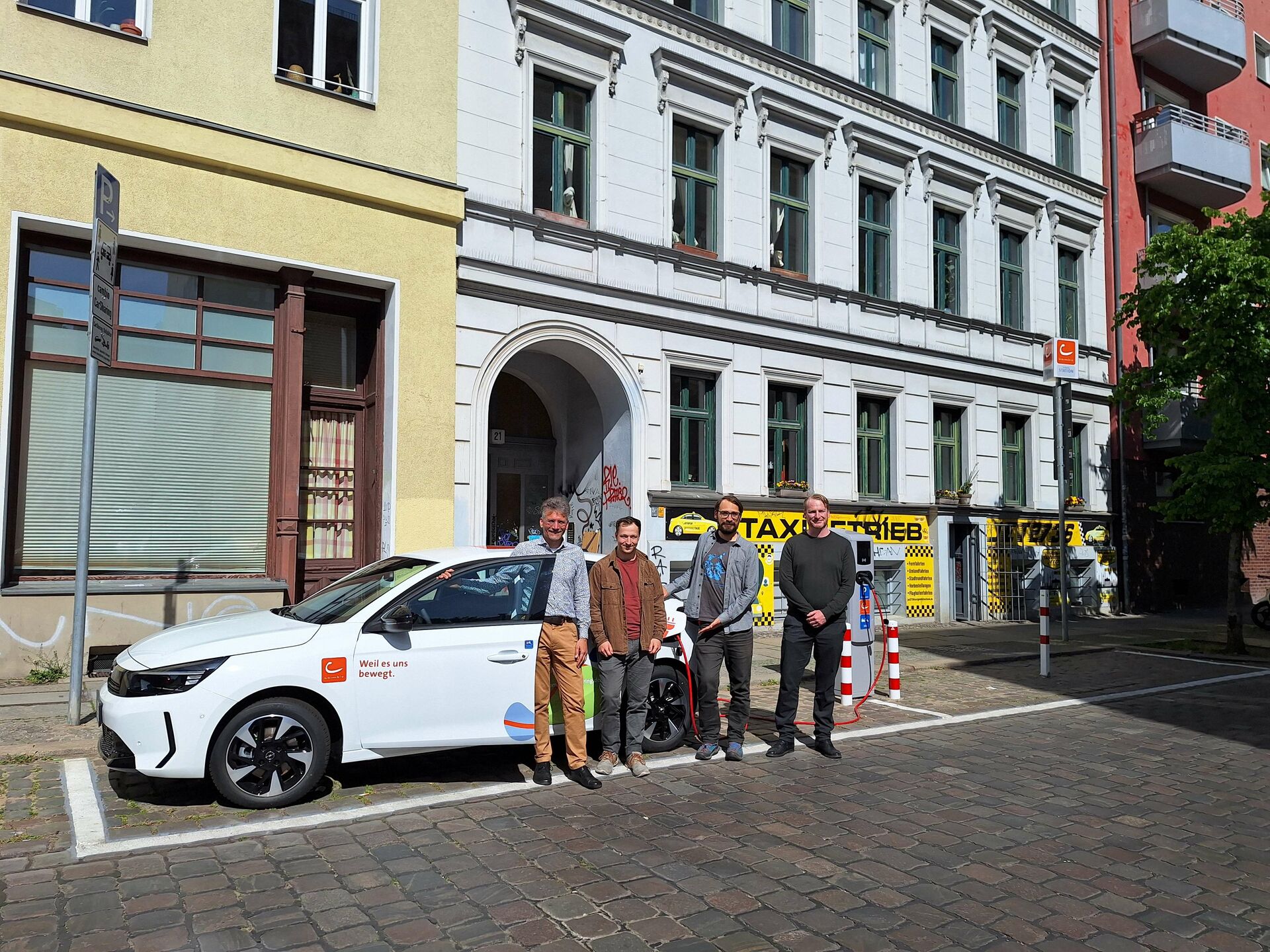 Vier Personen stehen vor einem weißen E-Auto, das an einer Ladesäule angeschlossen ist. Das Auto parkt am Straßenrand an einer CarSharing-Station. Im Hintergrund sind Berliner Mehrfamilienhäuser zu sehen.