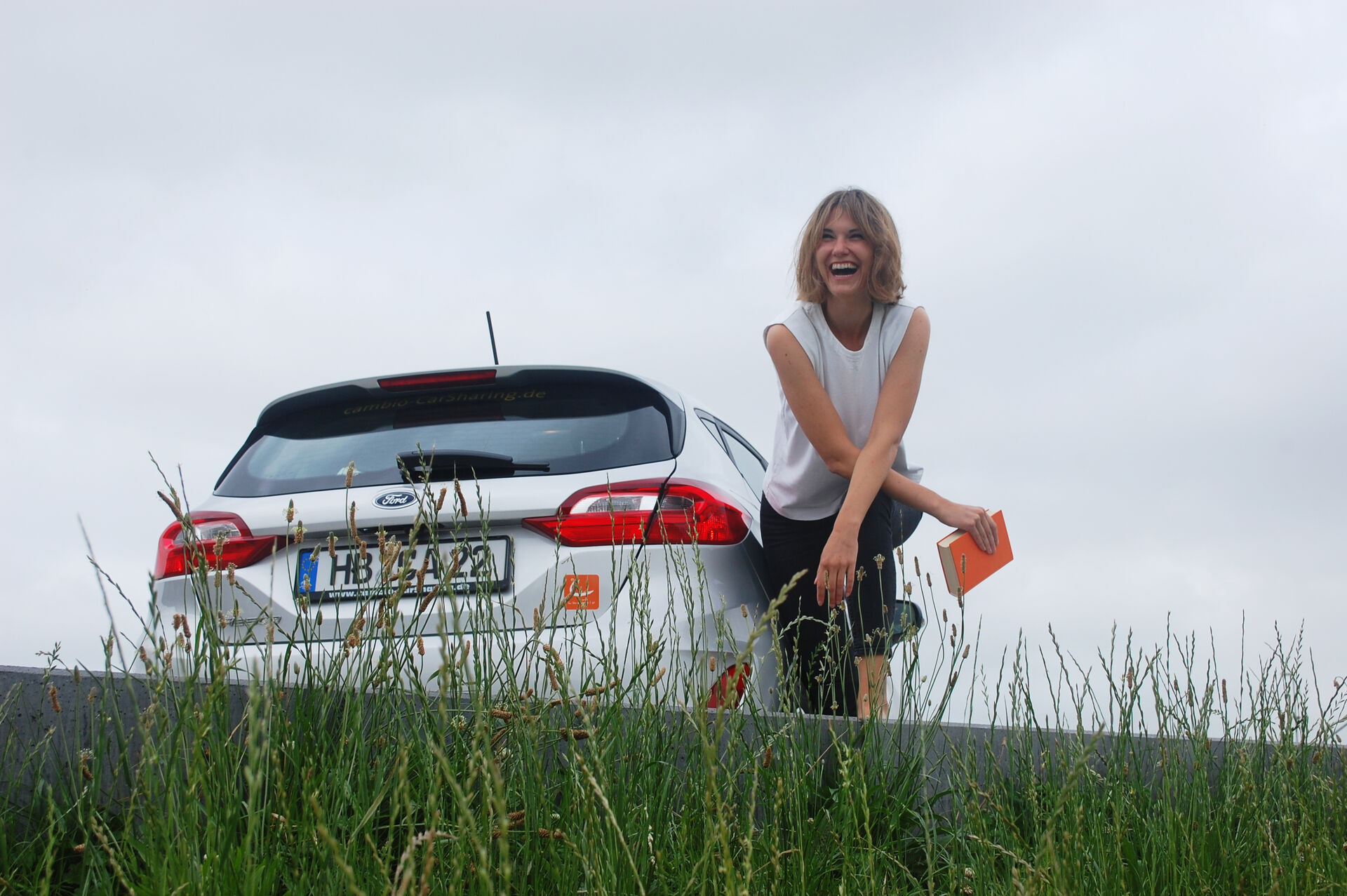 Rückansicht eines silberfarbenen Autos, daneben eine lachende Frau leicht vornübergebeugt mit einem orangenen Buch in der Hand. Beides ist leicht von hohen Gras bedeckt, im Hintergrund blaugrauer Himmel.