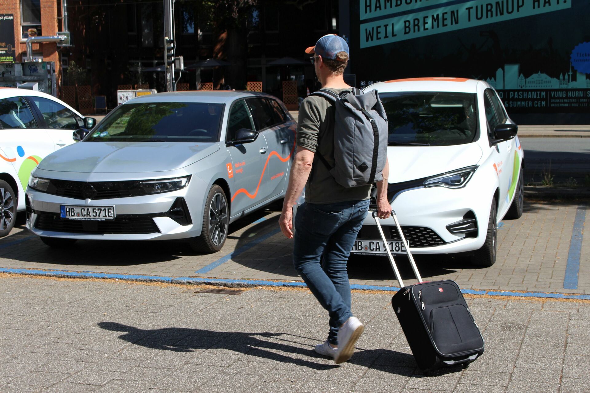 Ein Mann mit einem Rucksack und einem Koffer geht auf parkende E-Autos zu.