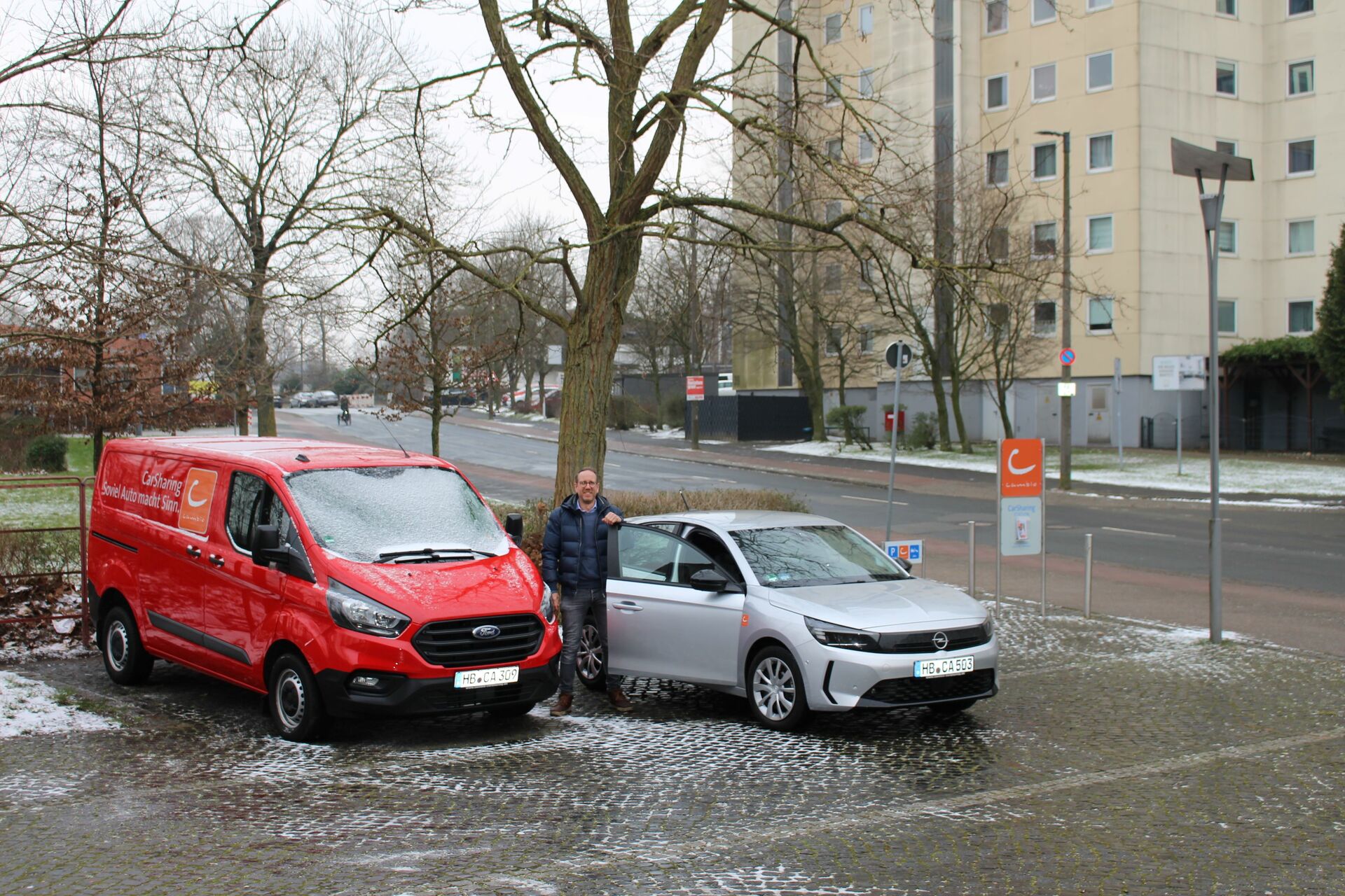 Eine Person steht zwischen einem roten Ford Transit und einem silbernen Opel Corsa. Es liegt etwas Schnee und im Hintergrund sind Bäume sowie ein hoher Wohnblock zu sehen. 