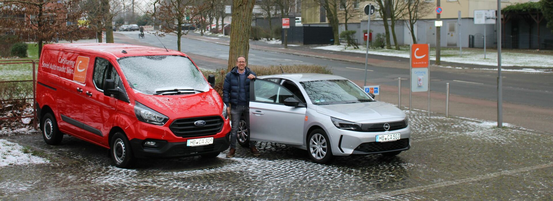 Eine Person steht zwischen einem roten Ford Transit und einem silbernen Opel Corsa. Es liegt etwas Schnee und im Hintergrund sind Bäume sowie ein hoher Wohnblock zu sehen. 