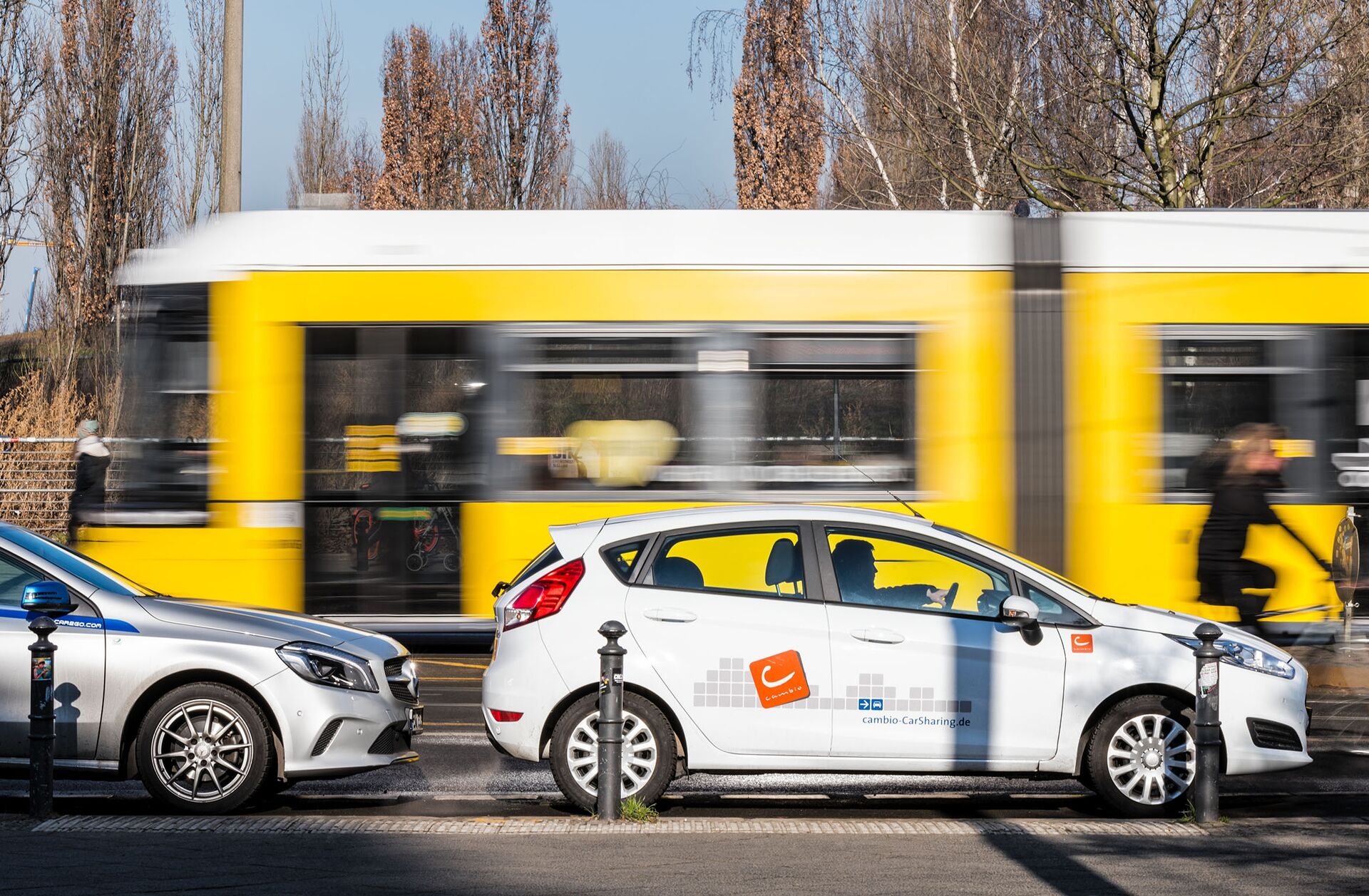 Weißes E-Auto von cambio steht auf der Straße, im Hintergrund fährt eine Straßenbahn vorbei.