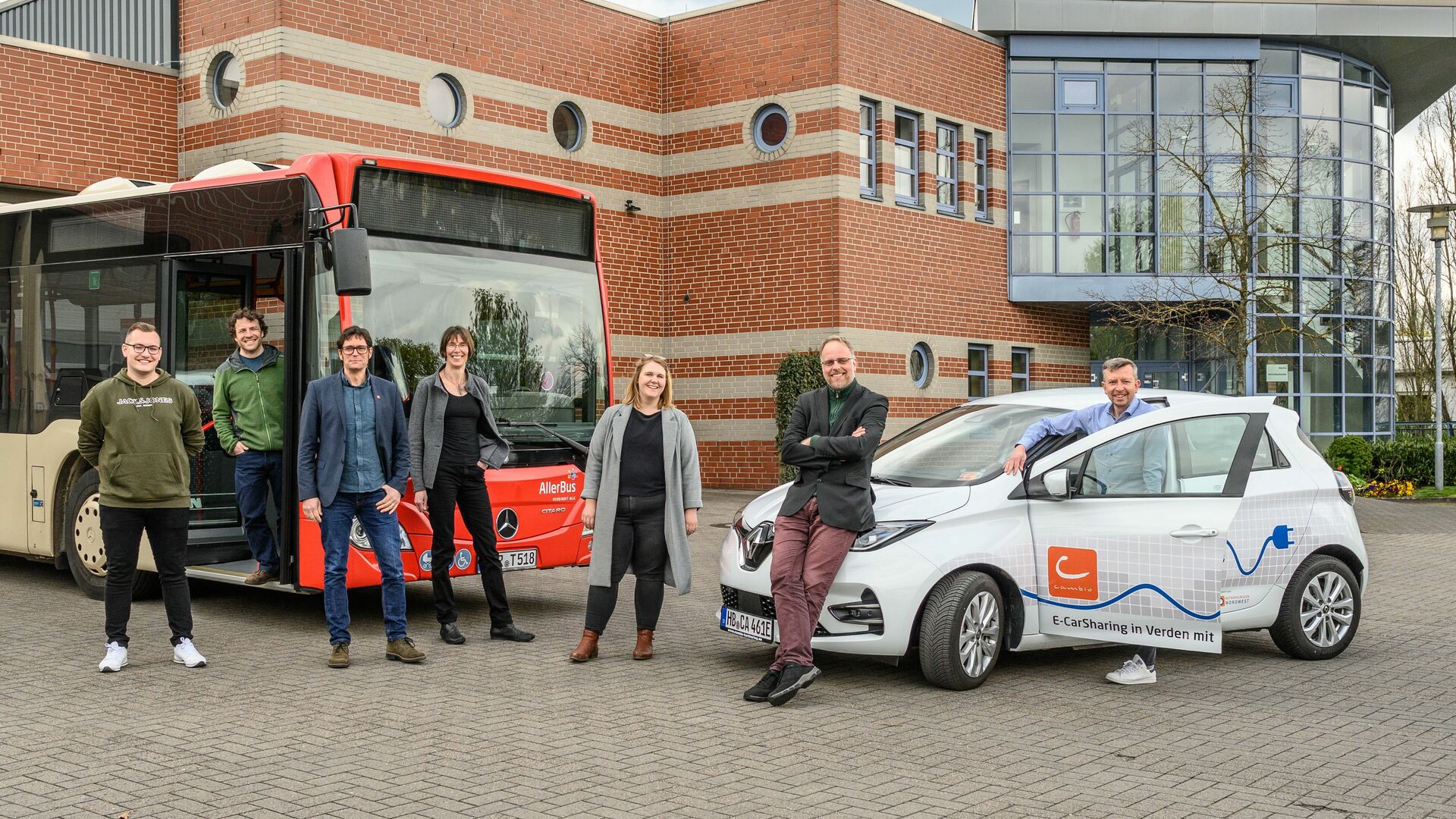 Eine Gruppe von Männern und Frauen stehen in Verden vor einem cambio-Auto und einem AllerBus.Mehrer Menschen stehen 