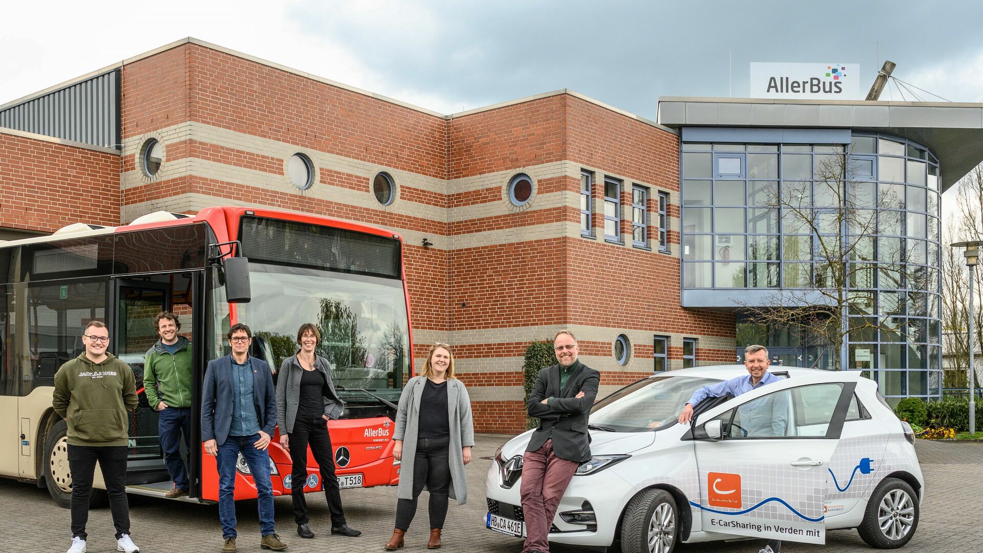 Eine Gruppe von Männern und Frauen stehen in Verden vor einem cambio-Auto und einem AllerBus.Mehrer Menschen stehen 