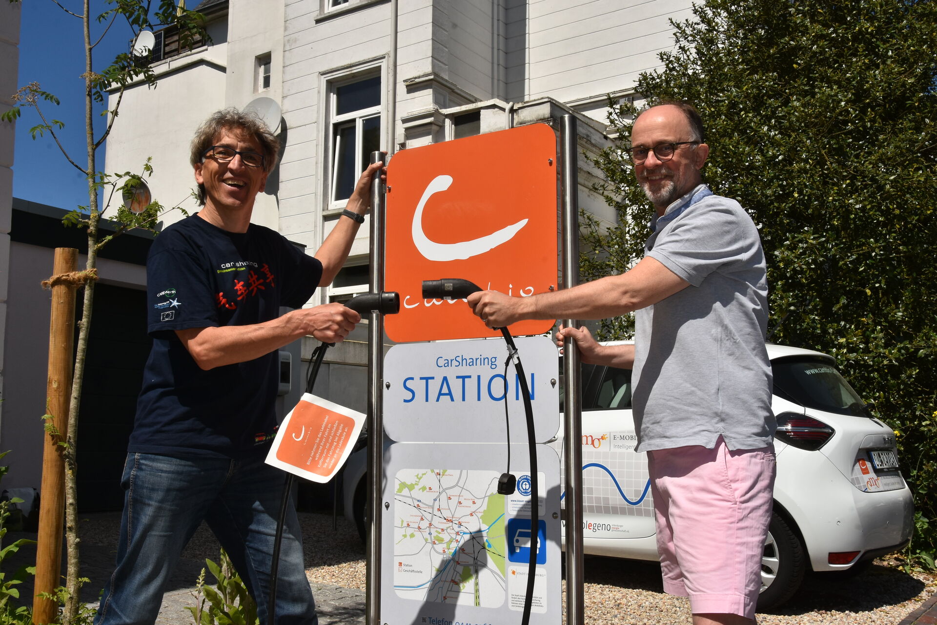 Zwei Personen stehen vor einem Stationsschild des CarSharing-Anbieters cambio. In der Hand halten sie je einen Ladestecker eines E-Autos. Dahinter parkt ein E-Auto. Im Hintergrund ist ein Haus zu sehen.