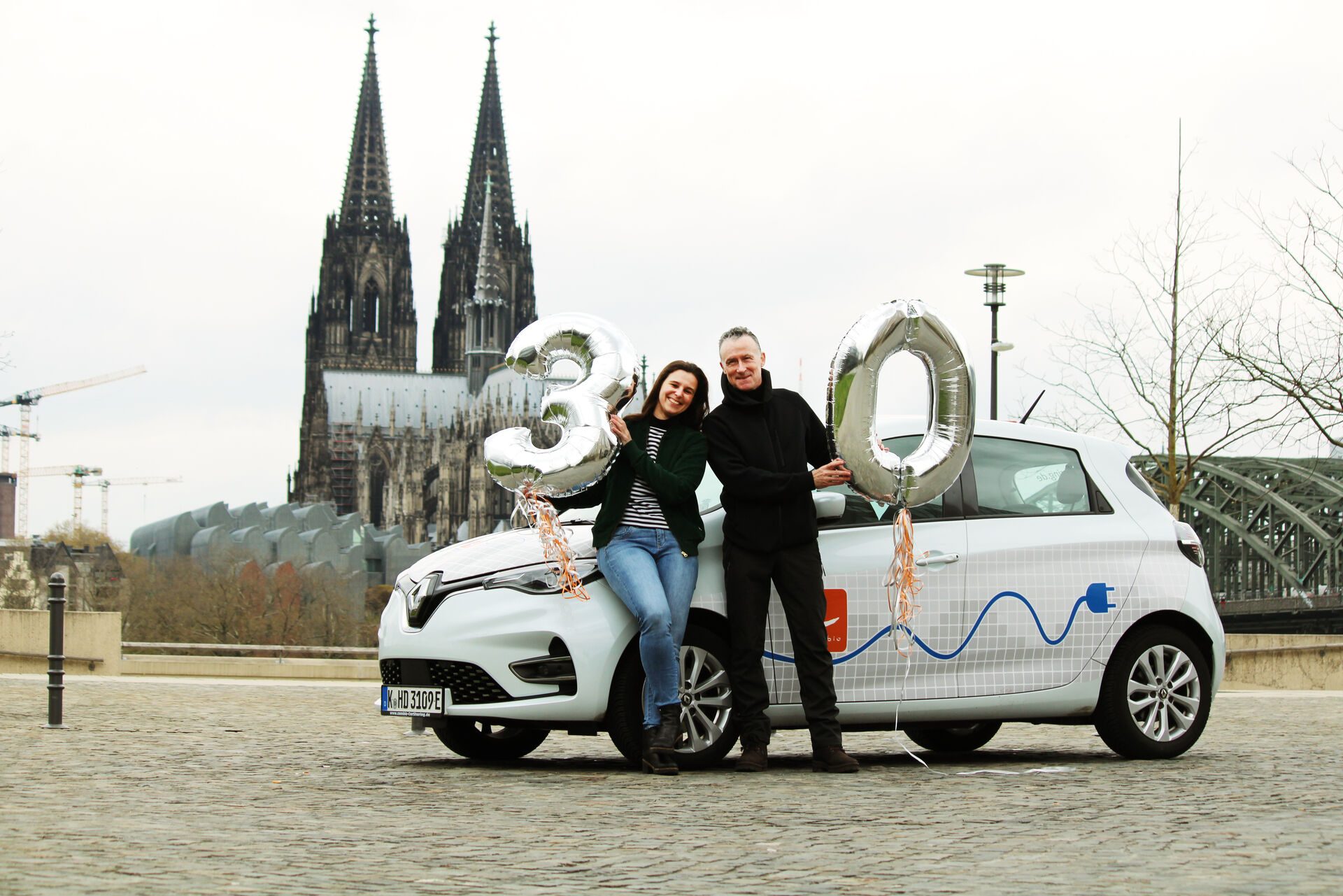 Mann und Frau stehen vor einem weißen Auto und halten eine Ballon 30 in die Kamera. Im Hintergrund sieht man den Kölner Dom.
