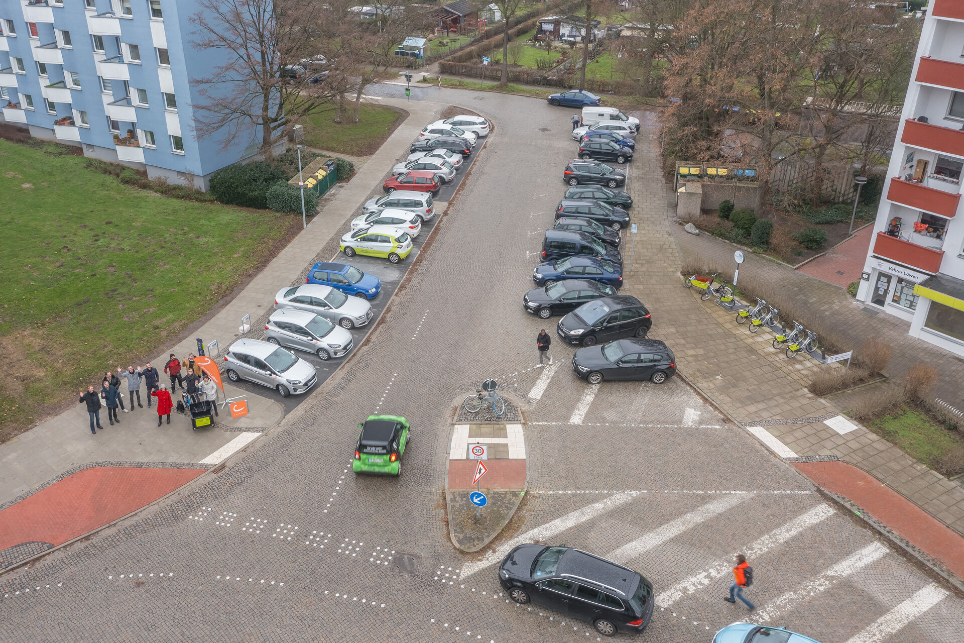 Eine Straße zwischen zwei Mehrfamilienhäusern, rechts und links neben der Straße stehen geparkte Autos, außerdem steht eine Gruppe von Leuten auf dem Fußgängerweg