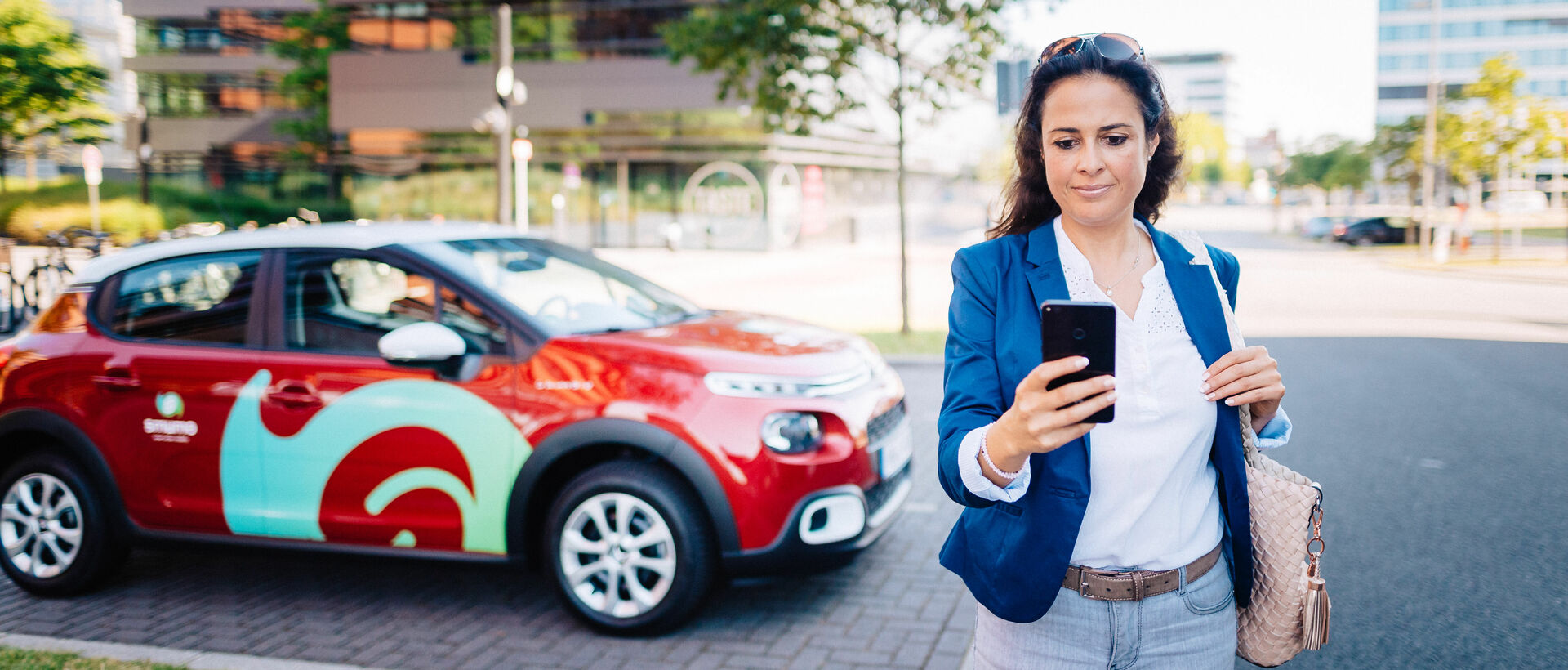 Im Vordergrund: Frau schaut auf Smartphone, im Hintergrund ein rotes smumo-Auto vor modernem Bürogebäude