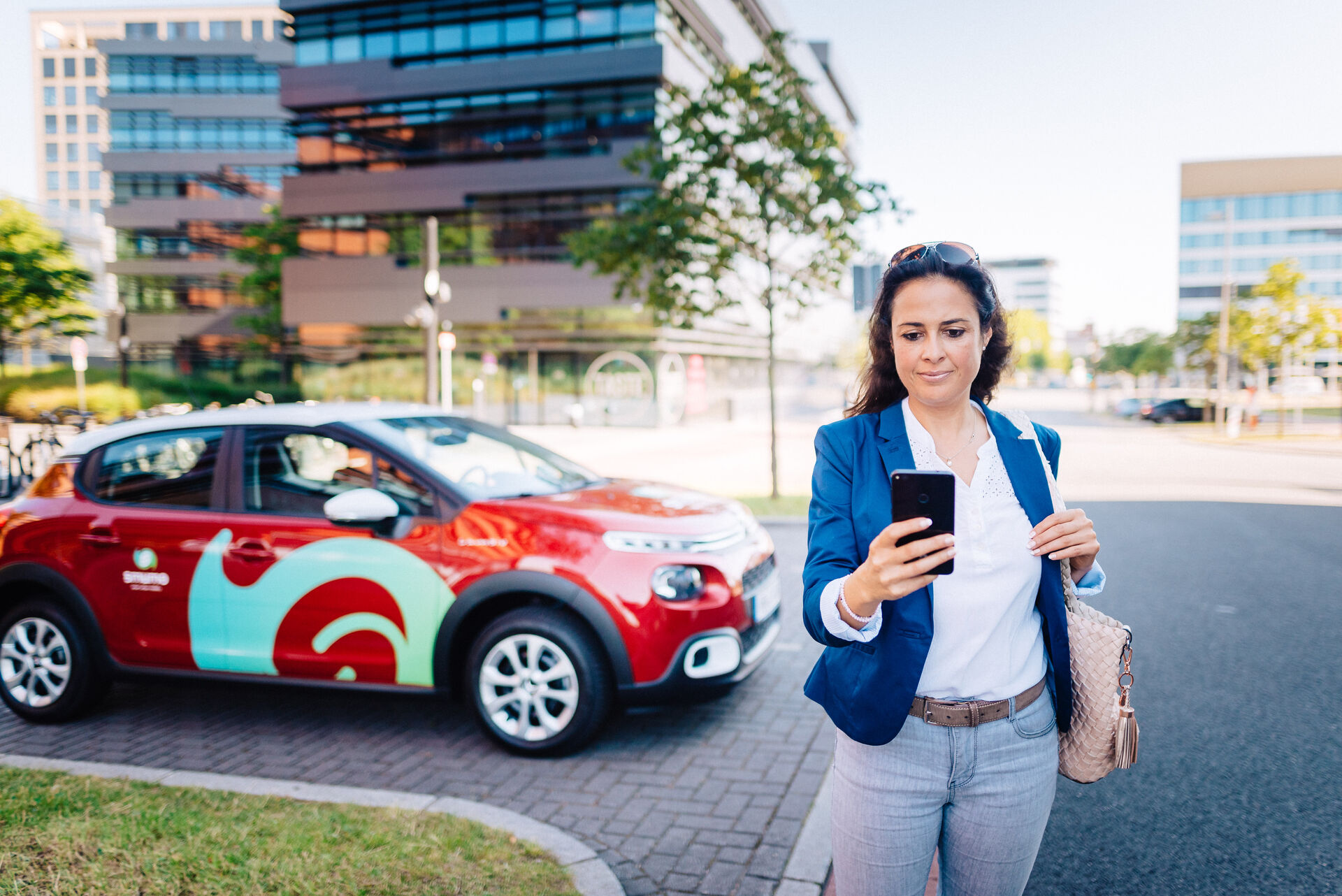 Im Vordergrund: Fau schaut auf Smartphone, im Hintergrund ein rotes smumo-Auto vor modernem Bürogebäude