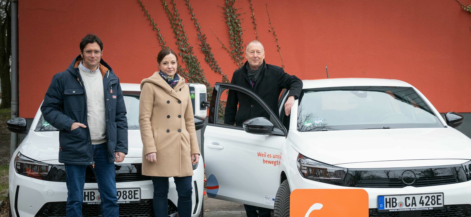 Drei Personen stehen vor zwei E-Autos vom Typ Opel Corsa-e. Im Vordergrund steht ein Schild mit dem Logo des CarSharing-Unternehmens cambio. Im Hintergrund befindet sich eine Mauer.