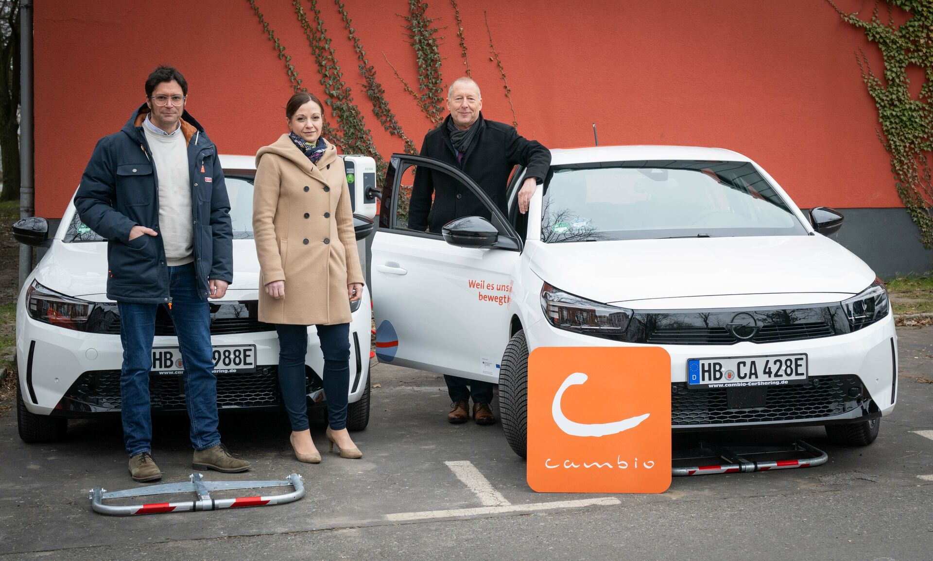 Drei Personen stehen vor zwei E-Autos vom Typ Opel Corsa-e. Im Vordergrund steht ein Schild mit dem Logo des CarSharing-Unternehmens cambio. Im Hintergrund befindet sich eine Mauer.