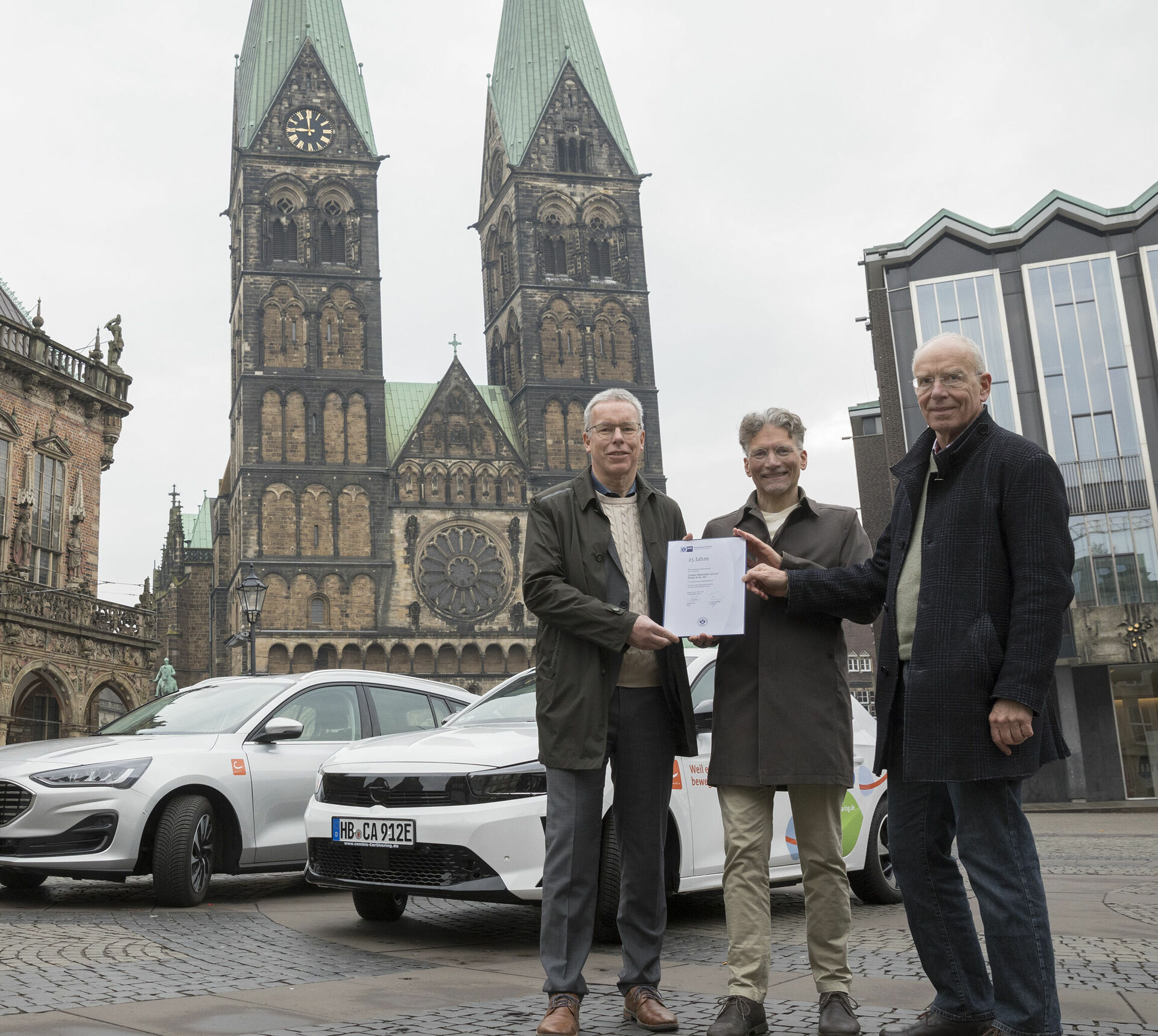 Drei Personen halten eine Urkunde in das Bild. Dahinter stehen ein weißes und ein silbernes Auto. Im Hintergrund ist das Bremer Rathaus, der Dom und die Bürgerschaft zu sehen.