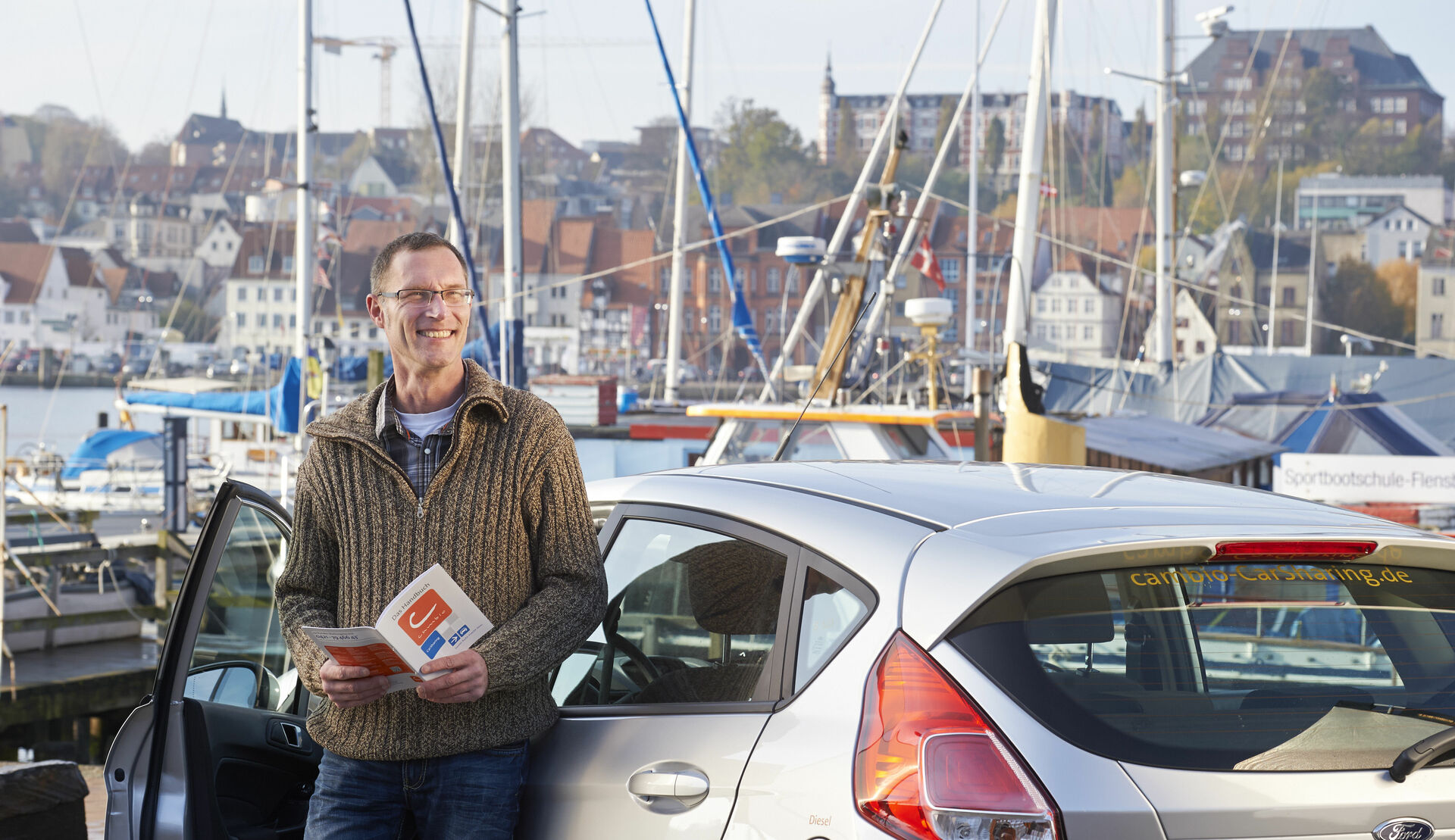 Mann steht mit einem cambio-Flyer lächelnd vor einem silbernen Auto. Im Hintergrund sieht man eine Hafenstadt
