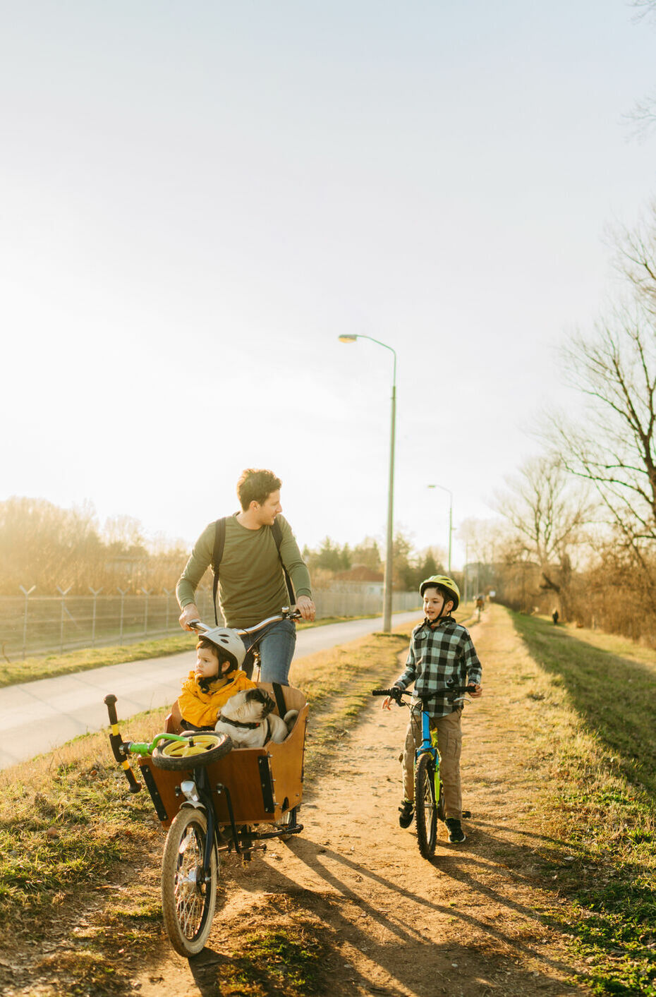 Ein Mann fährt ein Kind im Lastenrad herum und spricht mit einem weiteren Kind, das neben ihm auf einem Fahrrad fährt.
