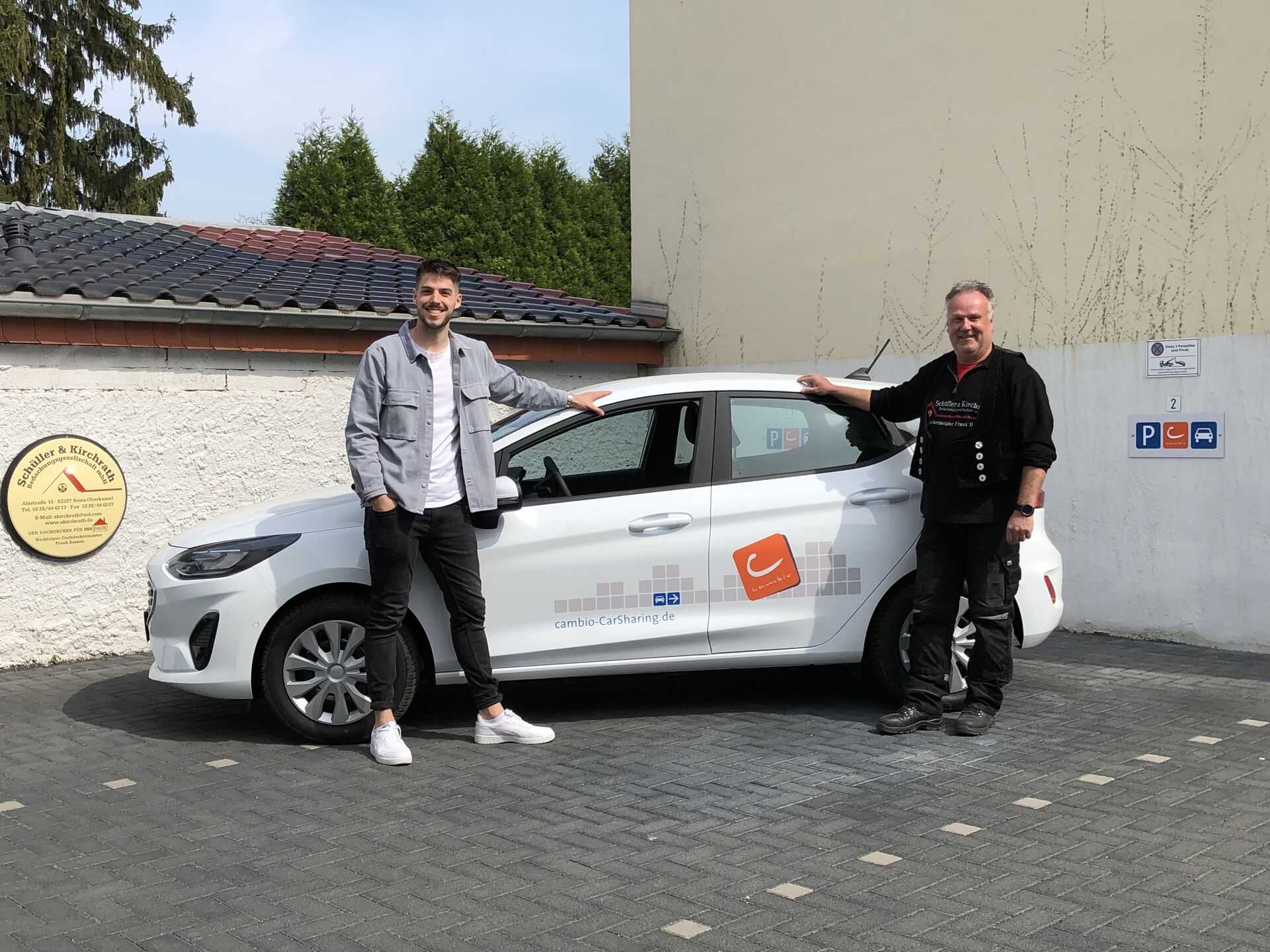 Zwei Männer stehen stolz vor einem weißen Auto mit einem Cambio Aufdruck.