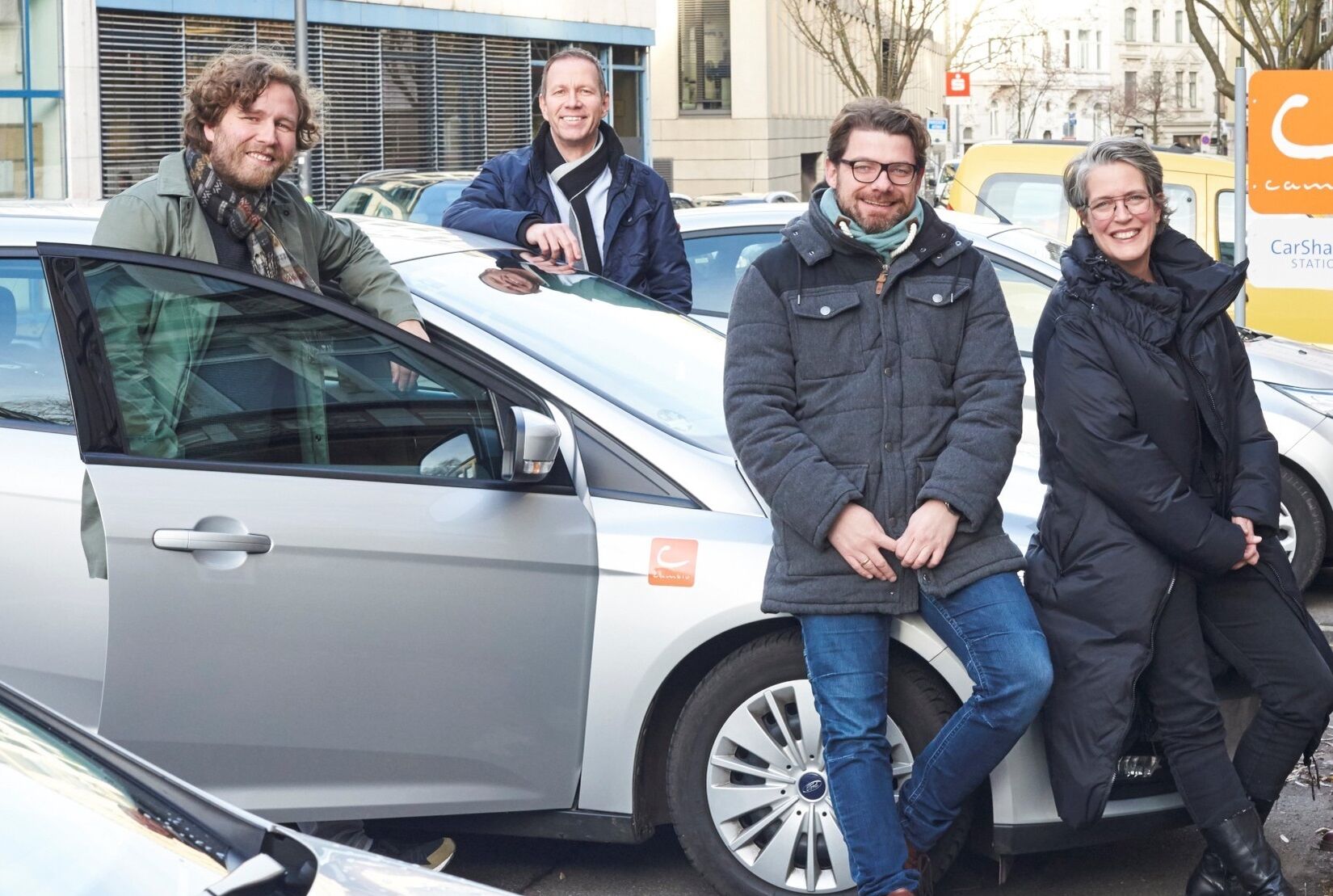 Vier Personen stehen an einem CarSharing-Auto und lächeln in die Kamera.