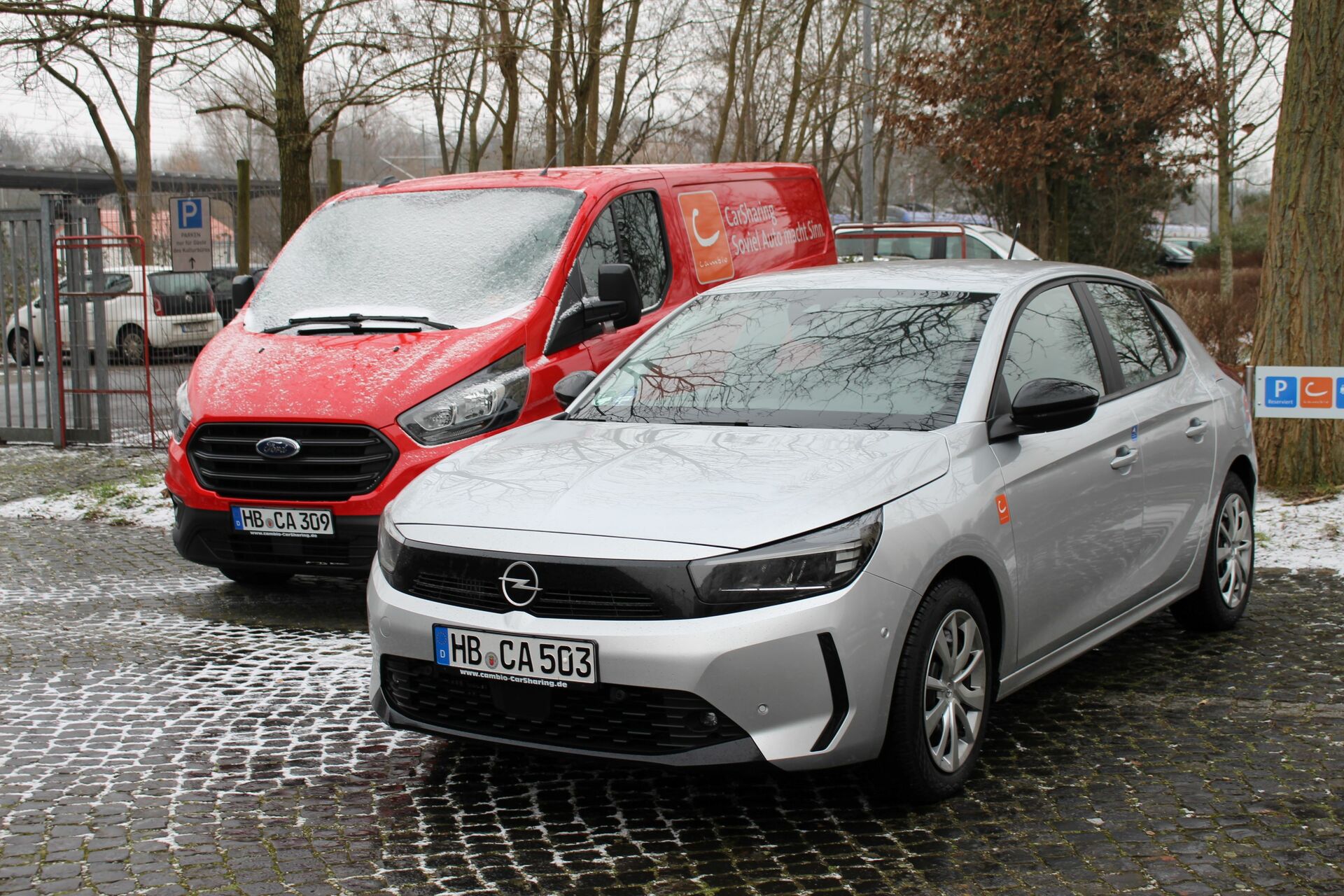 Ein silberner Opel Corsa steht vor einem roten Ford Transit Transporter. Auf den Fahrzeugen und dem Boden liegt etwas Schnee. Im Hintergrund stehen Bäume.