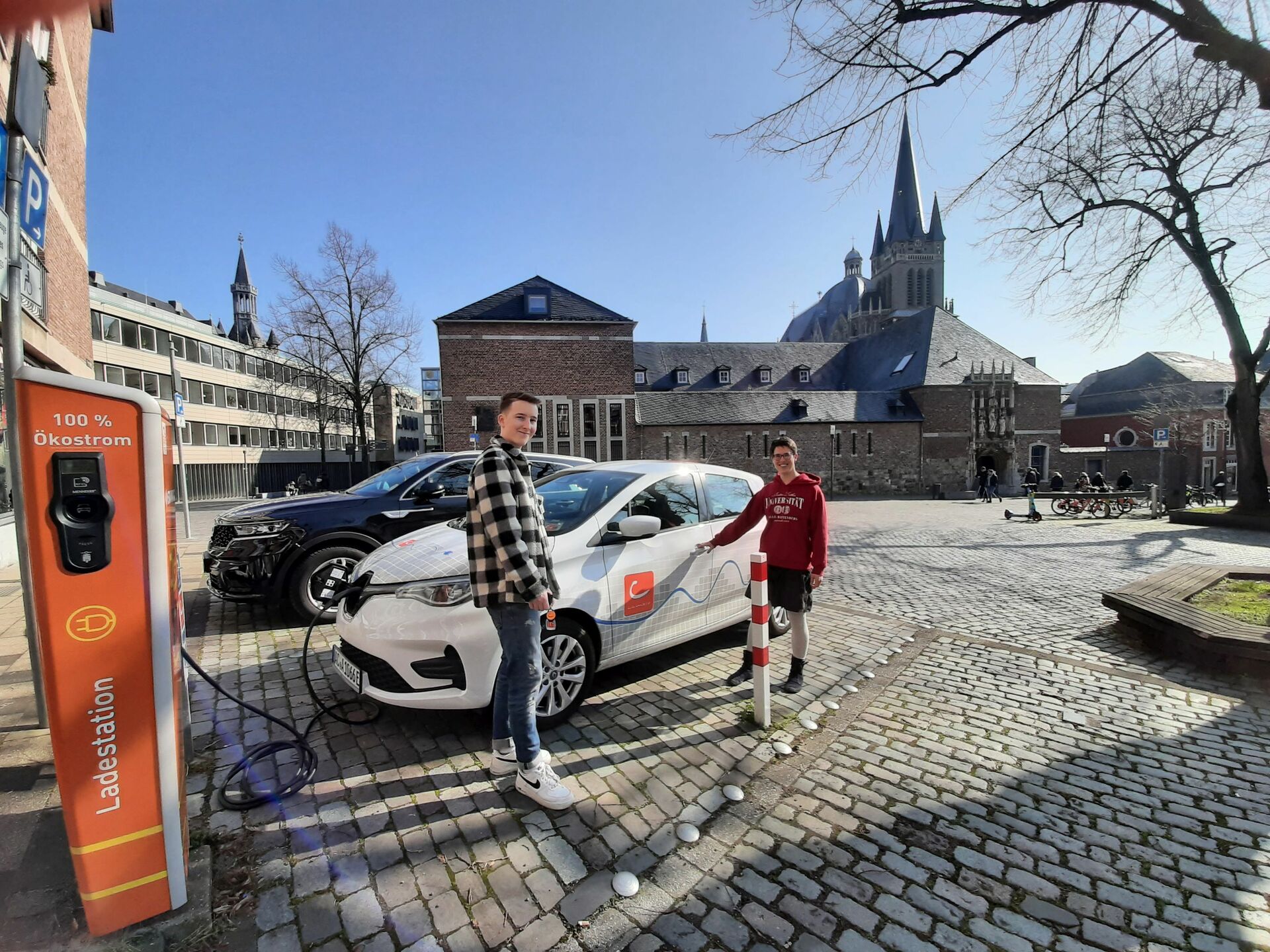 Zwei Männer stehen neben einem cambio-Auto an einer Ladesäule. Im Hintergrund der Aachener Dom.