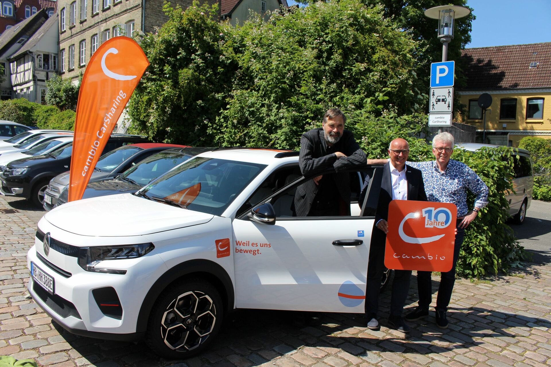 Drei Personen stehen an einem E-Auto. Das Fahrzeug steht auf einem Parkplatz. Im Hintergrund sind Büsche und Häuser zu sehen.