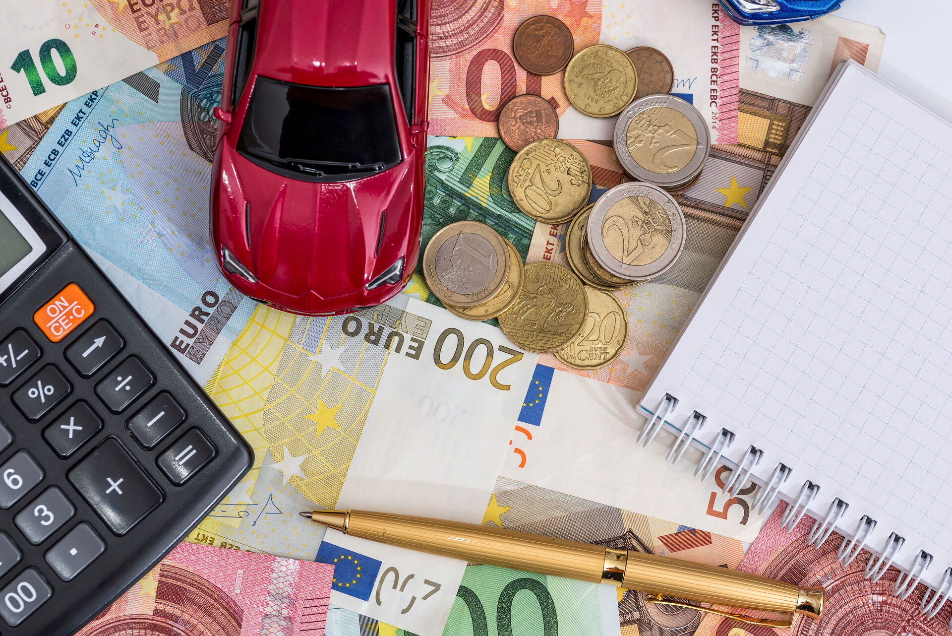 Auf einem Tisch liegen Euroscheine und Eurogeldstücke verstreut. Darauf ein Block und ein Stift, ein Taschenrechner und ein Spielzeugauto.