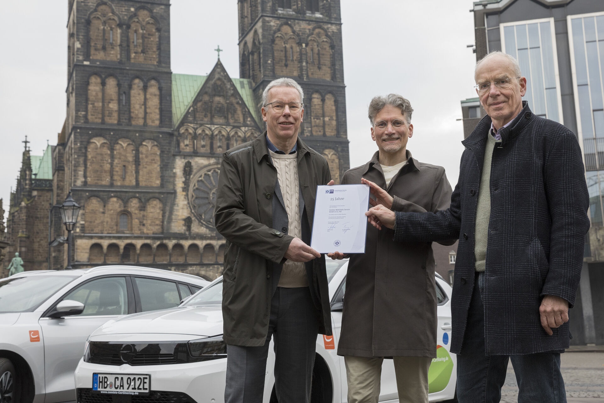 Drei Personen halten eine Urkunde in das Bild. Dahinter stehen ein weißes und ein silbernes Auto. Im Hintergrund ist das Bremer Rathaus, der Dom und die Bürgerschaft zu sehen.