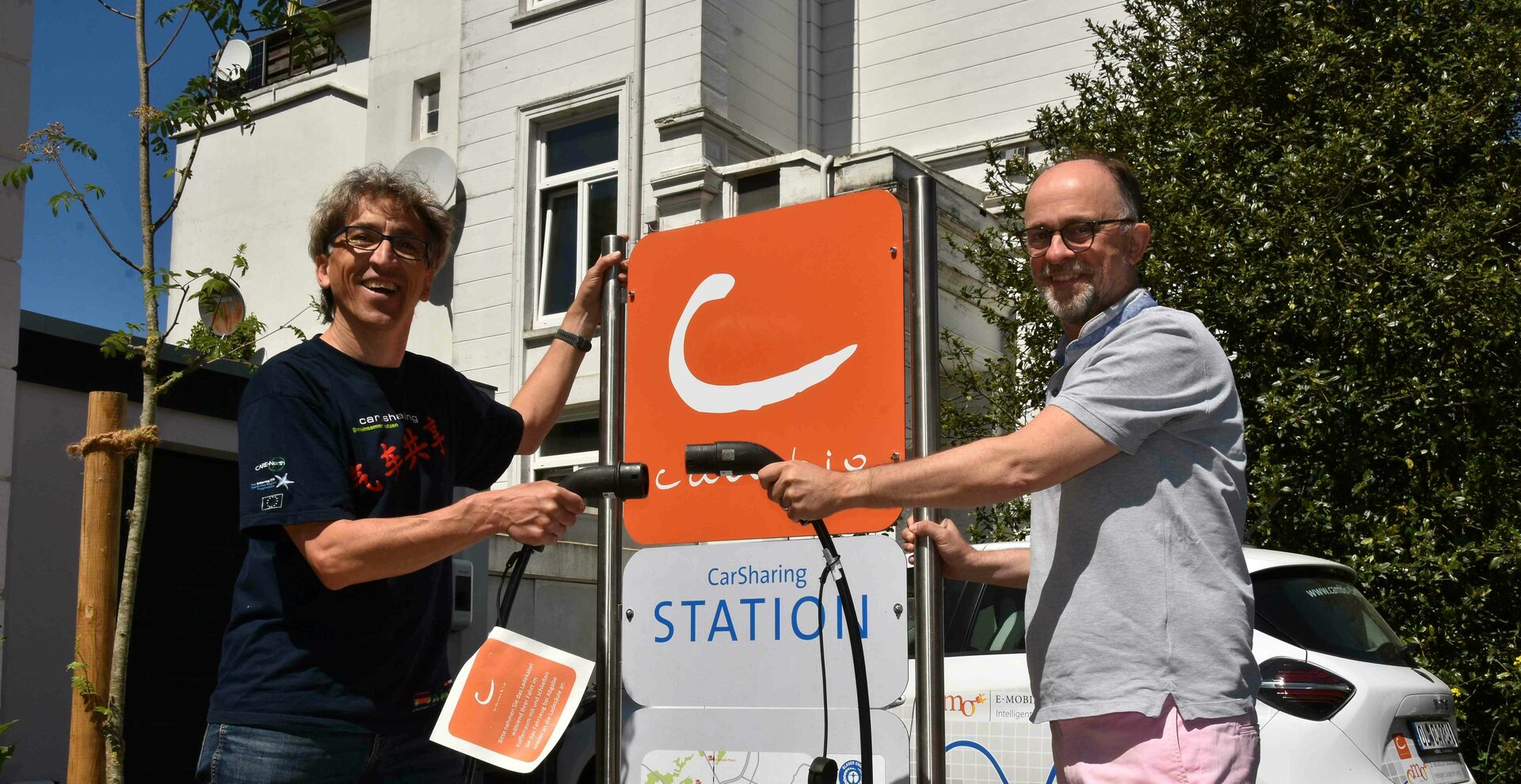 Zwei Personen stehen vor einem Stationsschild des CarSharing-Anbieters cambio. In der Hand halten sie je einen Ladestecker eines E-Autos. Dahinter parkt ein E-Auto. Im Hintergrund ist ein Haus zu sehen.