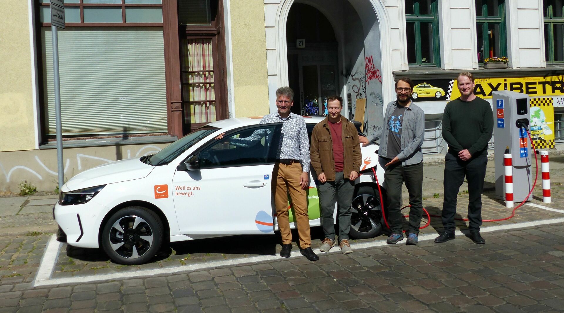 Vier Personen stehen vor einem weißen E-Auto und einer Ladesäule. Im Hintergrund sind Hausfassaden zu sehen.