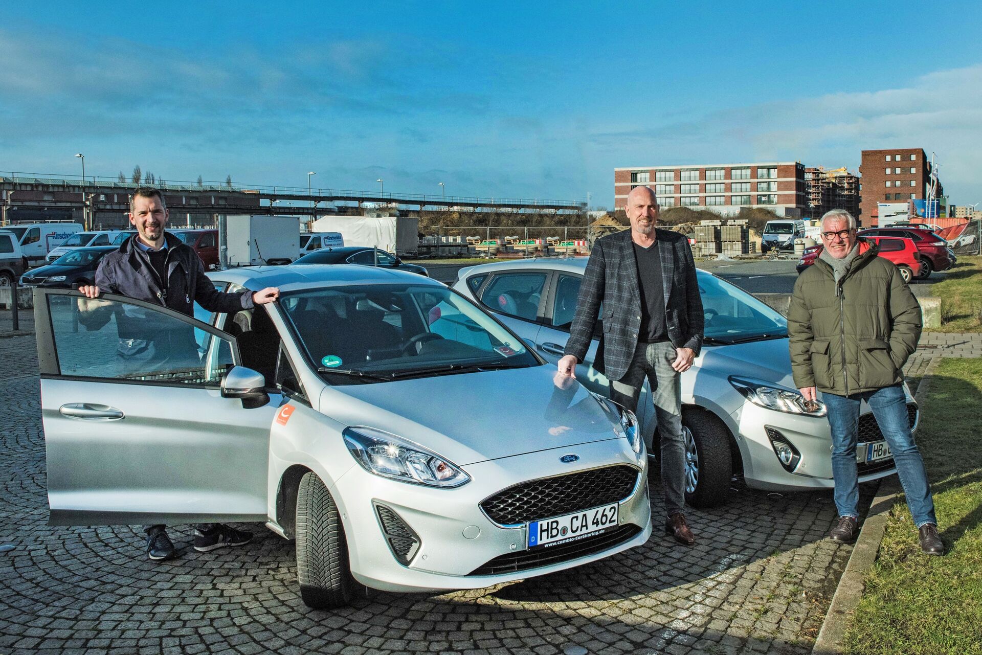 Drei Personen stehen neben zwei silbernen Autos