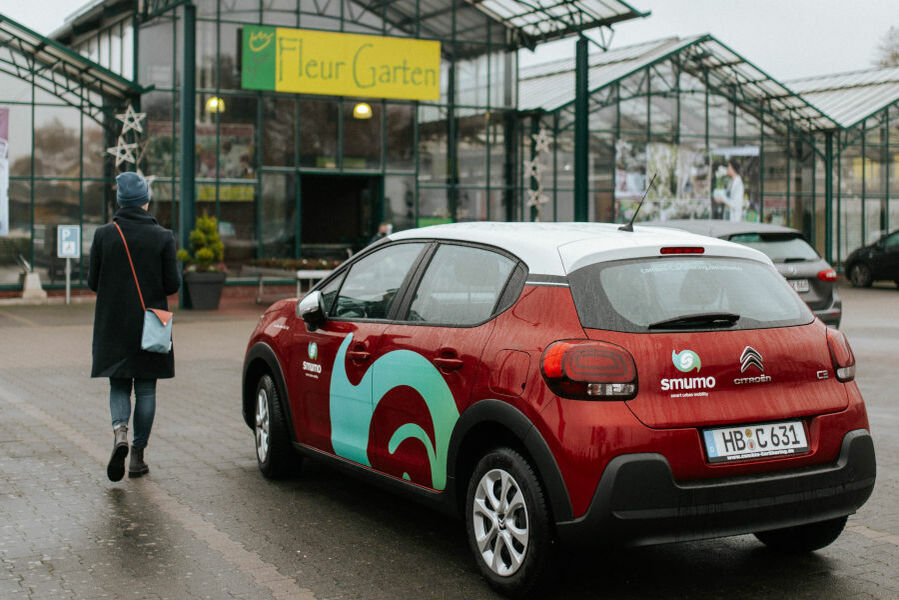 smumo-Auto daneben die Frau, Rückenansicht. Sie geht zum im Hintergrund gelegenen Gartencenter. Das Wetter ist regnerisch und grau.