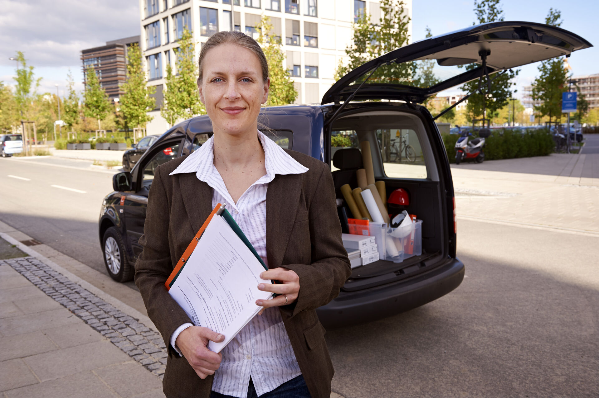 Frau steht vor einem Auto mit geöffneten Kofferraum. Darin sind mehrere Kisten und Papierrollen. Sie trägt Bluse und Blazer, schaut in die Kamera mit einem Lächeln. Ihre Haare sind zum Zopf gebunden. In der Hand hat sie Papiere, die ein Vertrag sein könnten.