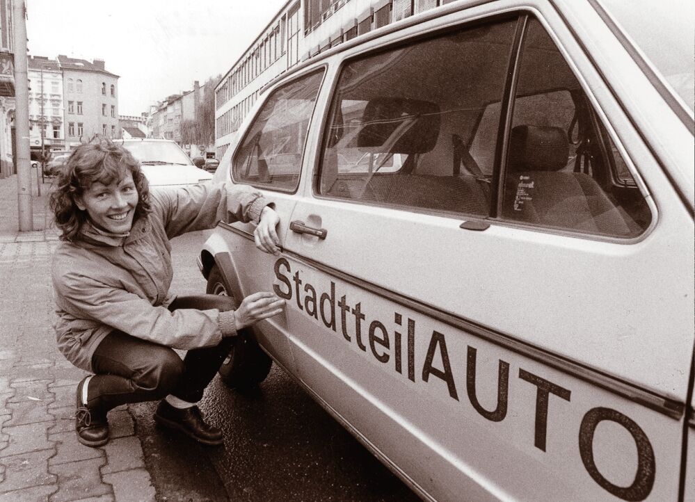 Ein schwarz-weiß-Bild, eine Frau kniet vor der Seite eines Autos und zieht dort den Kleber der Aufschrift "Stadtteil-Auto" ab.