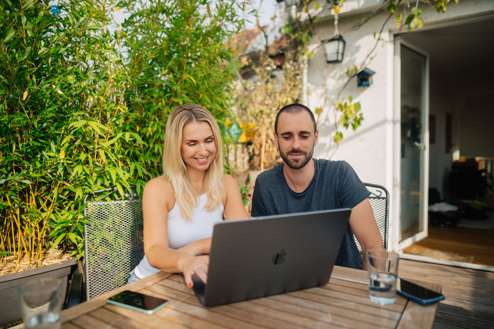 Eine Frau und ein Mann sitzen auf einer Terrasse an einem Gartentisch und gucken auf einen Laptop