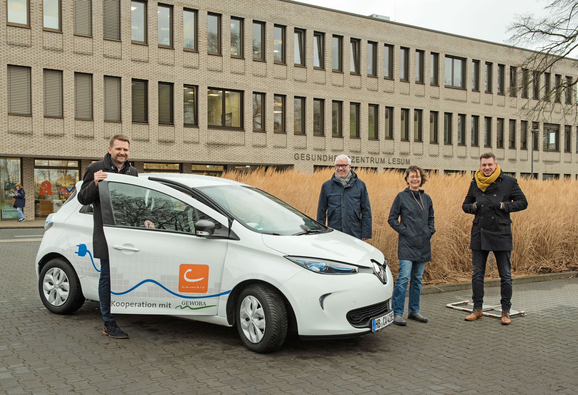 Ein Mann hält die Tür eins weißen E-Autos, rechts daneben stehen drei Personen und gucken fröhlich in die Kamera.