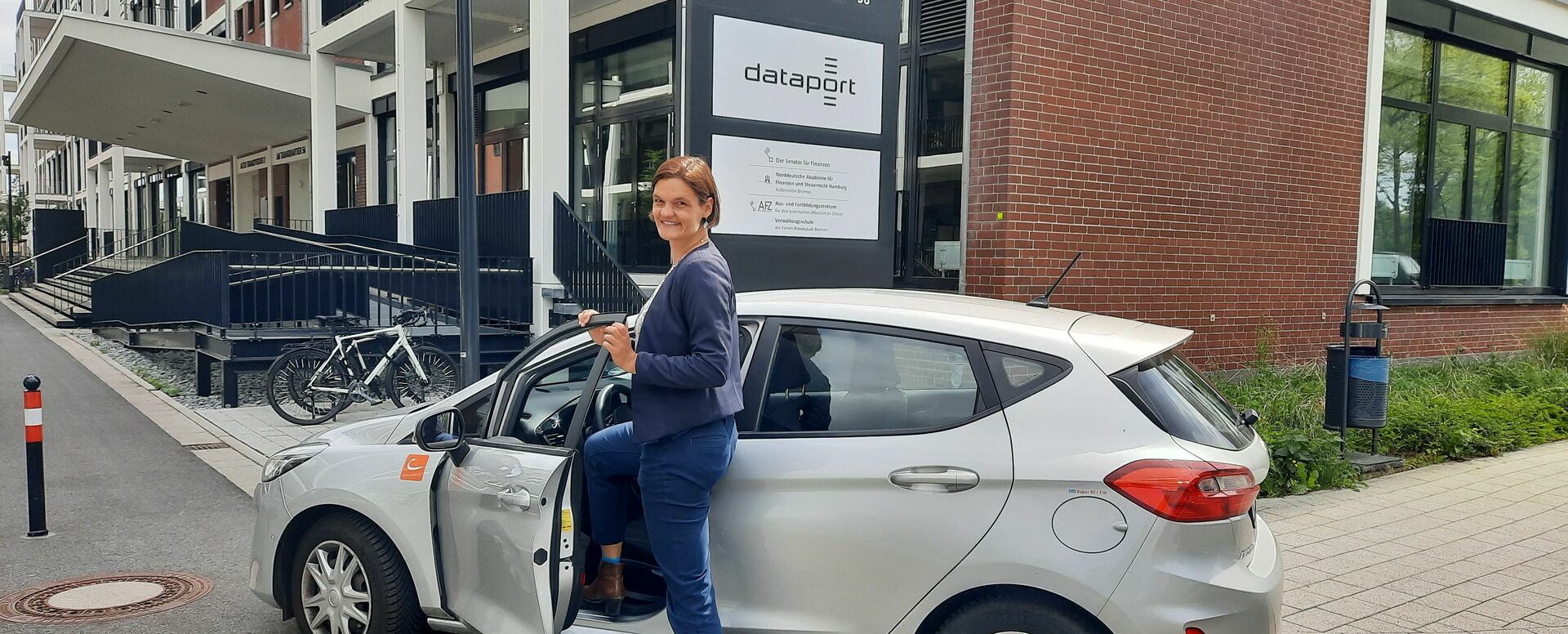 Eine Frau steigt in einen Kleinwagen ein und lächelt in die Kamera. Im Hintergrund ist ein Bürogebäude zu sehen.