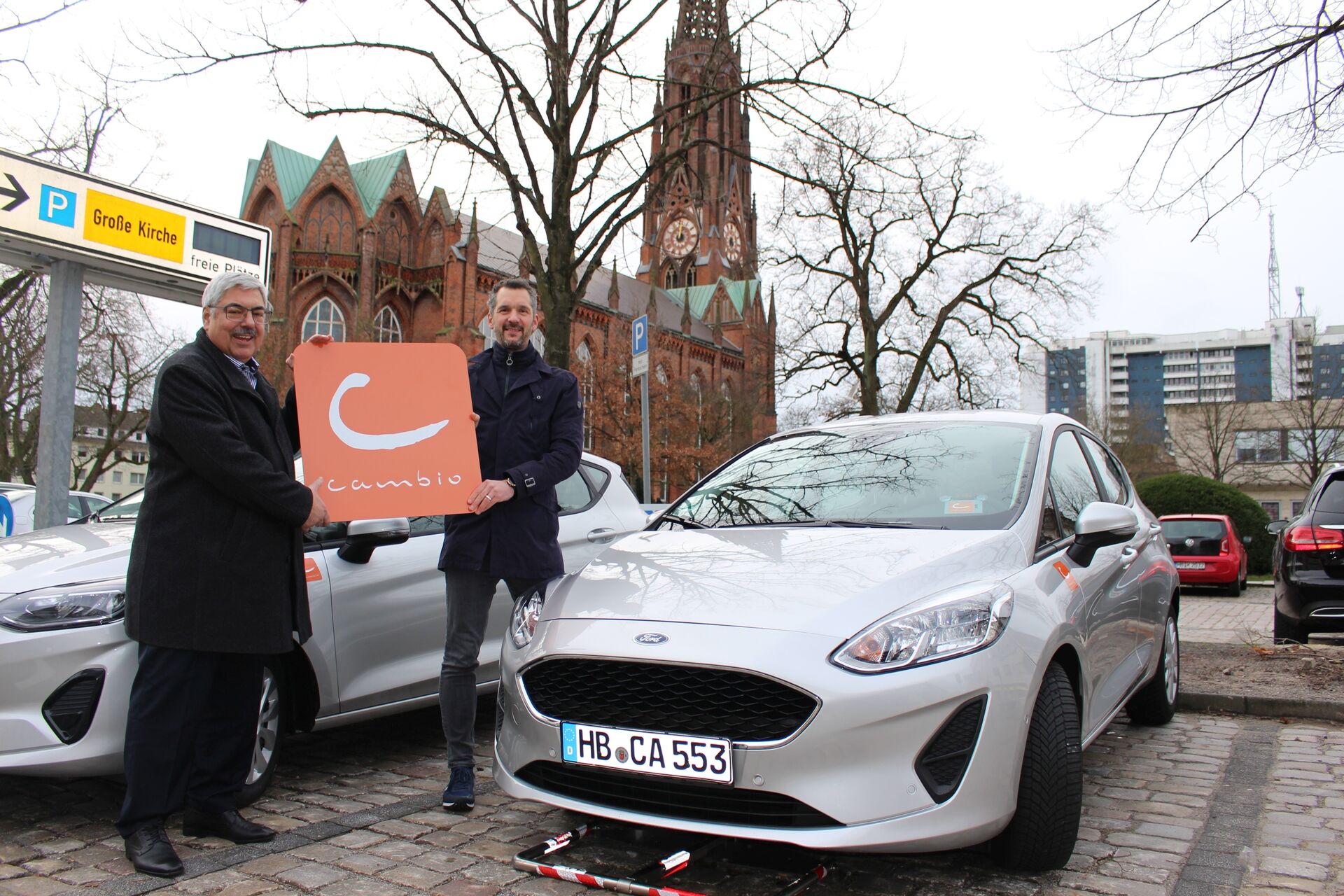 Zwei Männer stehen neben einem silbernen Auto und halten ein Cambio Schild hoch. Im Hintergrund sieht man eine Kirche. 