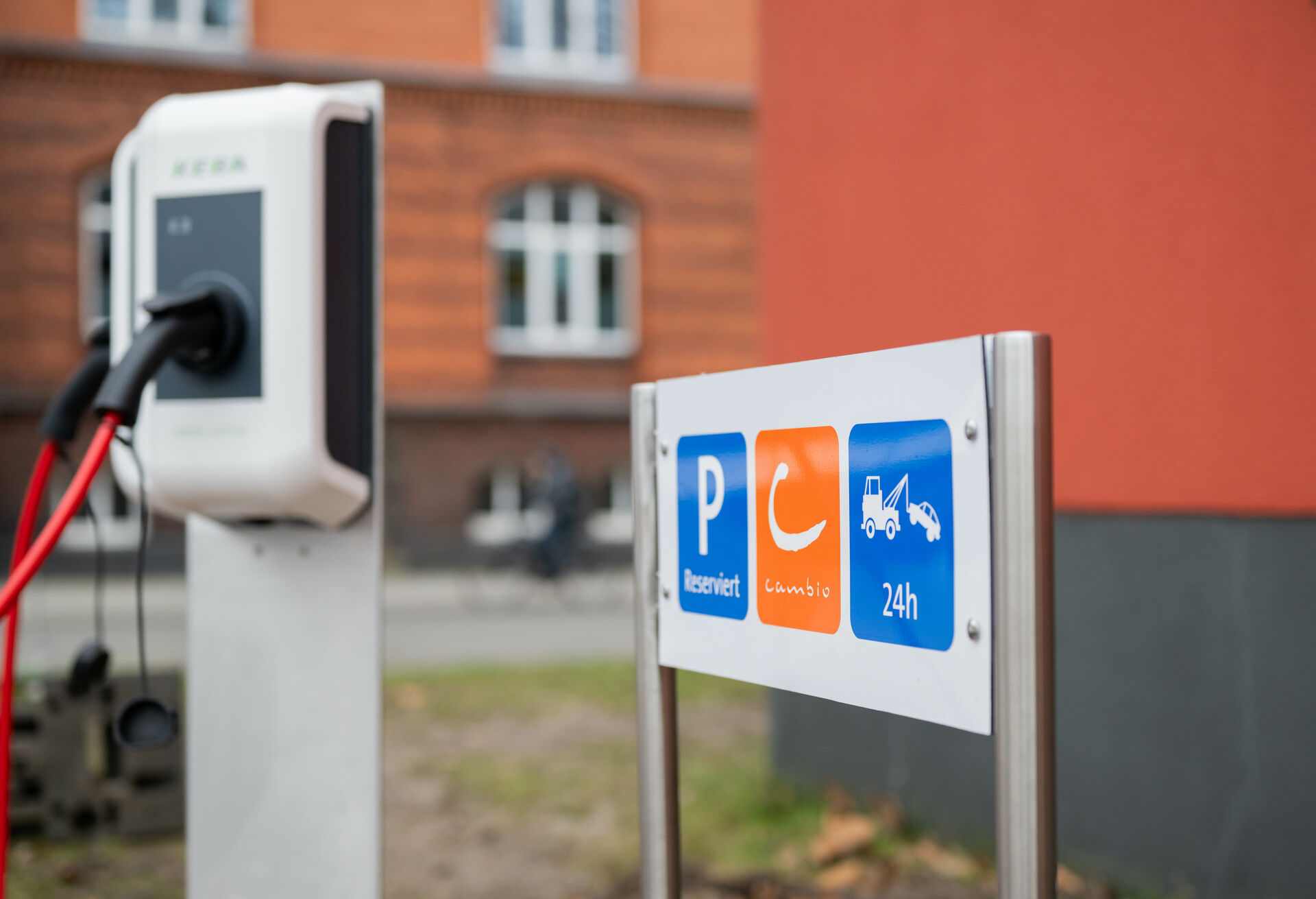 Ein Schild, auf dem "Reserviert", "cambio" und "24h" steht. Daneben ist eine Ladesäule für E-Autos zu sehen. Im Hintergrund fährt ein Fahrrad auf der Straße vor einem Gebäude.