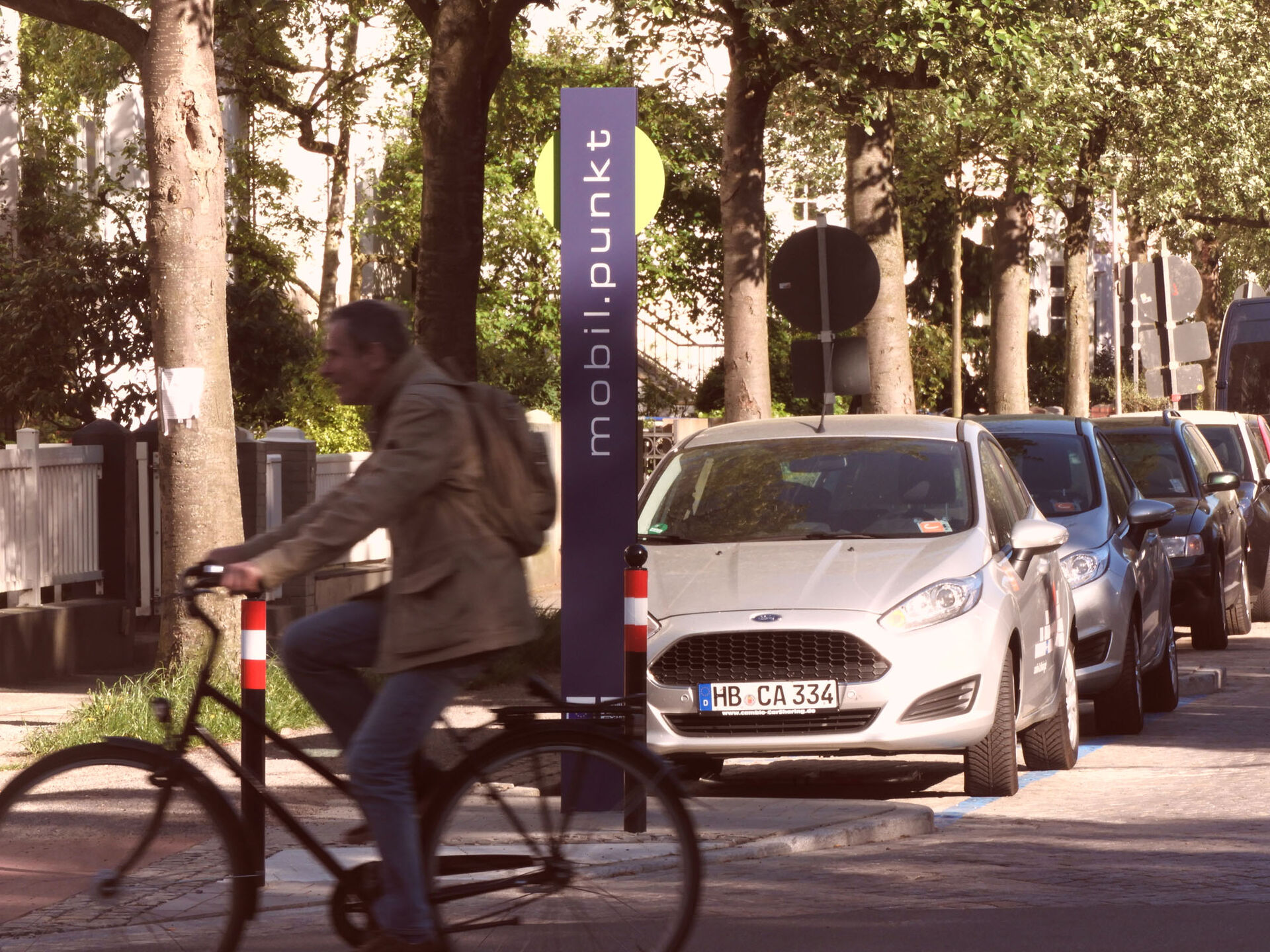 Im Vordergrund: Eine Person auf einem Fahrrad. Im Hintergrund: Ein parkendes cambio-Auto. Dazwischen: Ein Schild mit der Aufschrift mobilpunkt