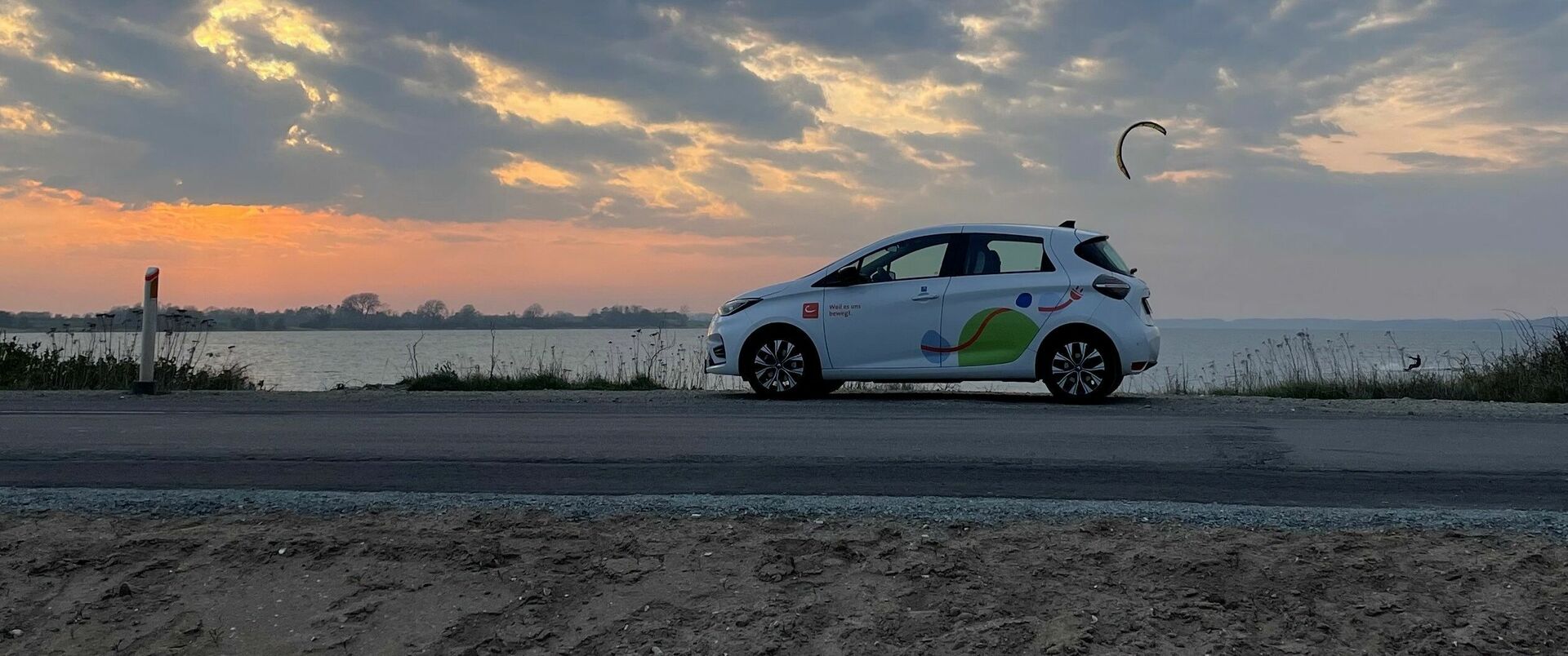 Ein E-Auto von cambio steht auf einer Straße. Im Hintergrund ist Wasser zu sehen, auf dem ein Kitesurfer unterwegs ist. Dahinter geht die Sonne unter.