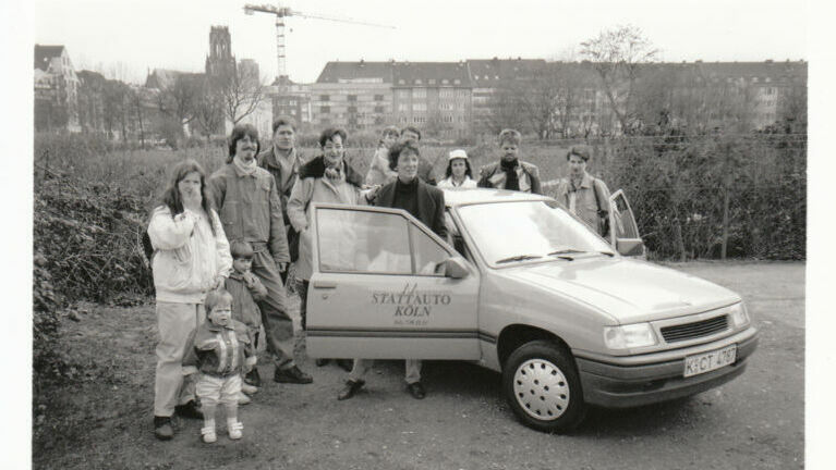 Das Bild ist in s/w es stehen mehrere Personen (von alt bis jung) neben dem Auto. Die Türen sind geöffnet.