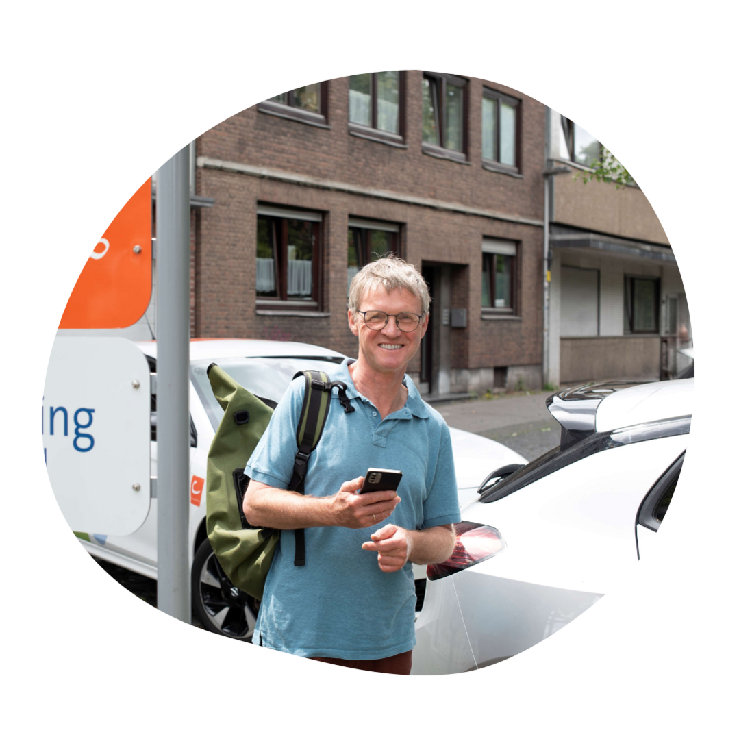 Ein Mann steht an einer CarSharing-Station und lächelt mit Smartphone in der Hand in die Kamera
