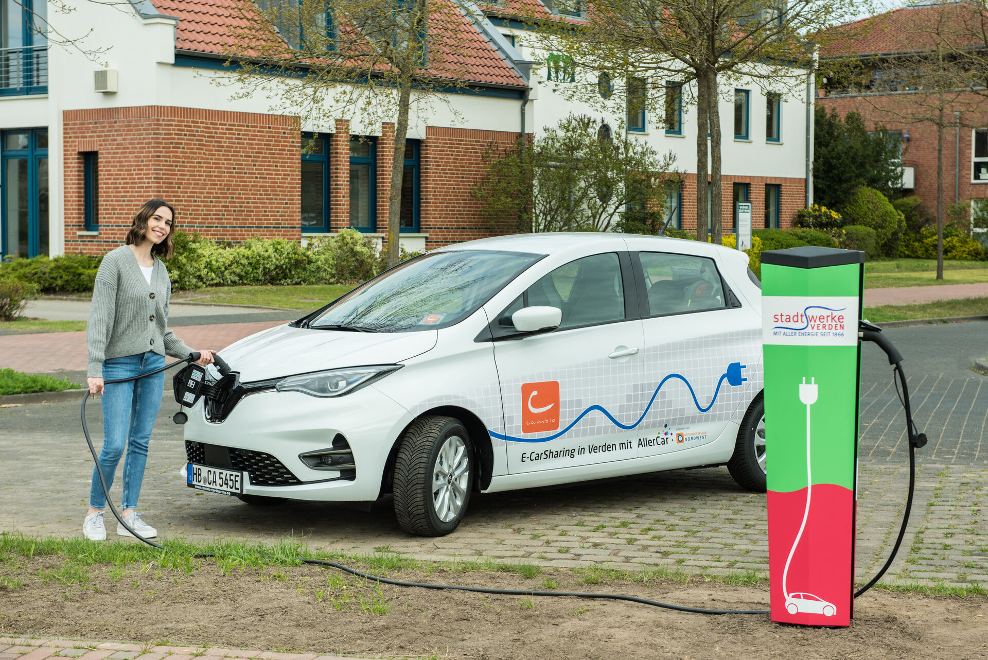 Eine Frau steht lächelnd vor einem E-Auto und schließt es an die LadesäuleZapfsäule an.