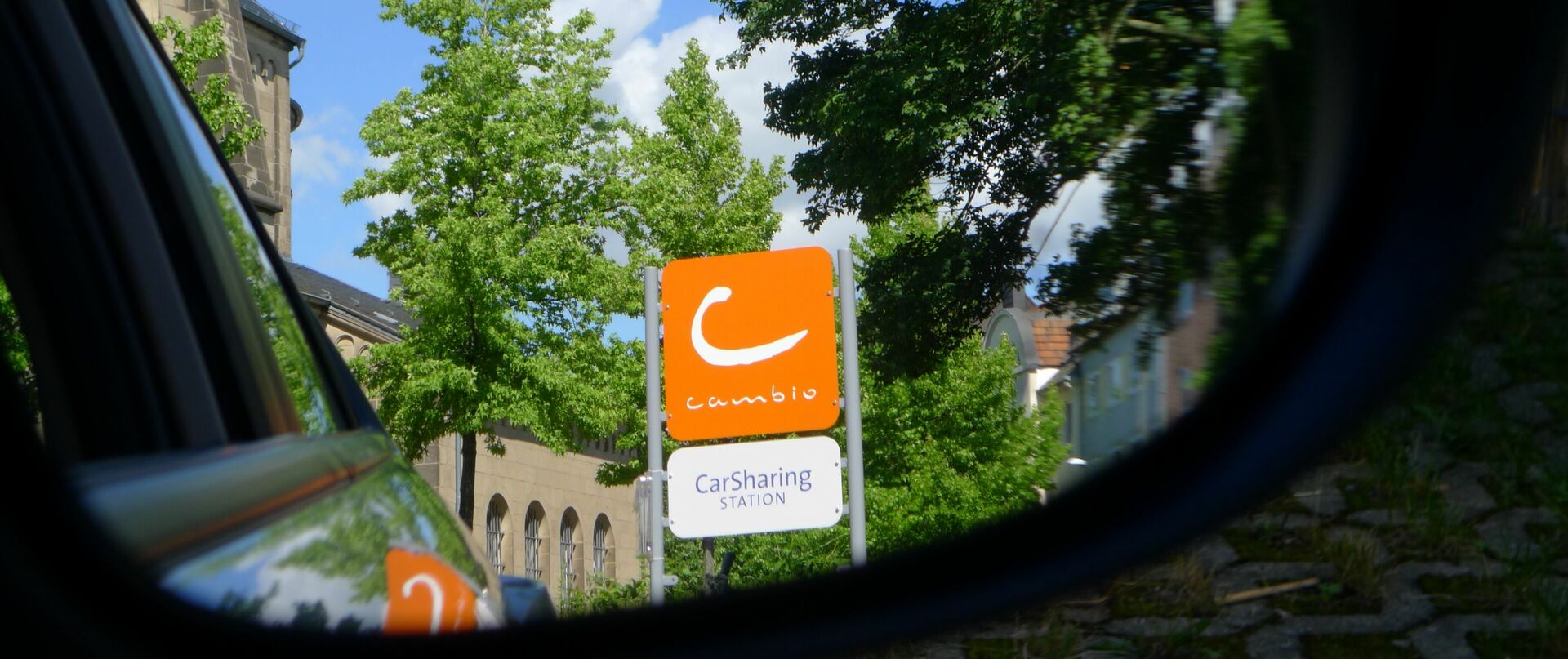 Nahaufnahme eines Autorückspiegels, in dem ein Stationsschild von cambio zu sehen ist 