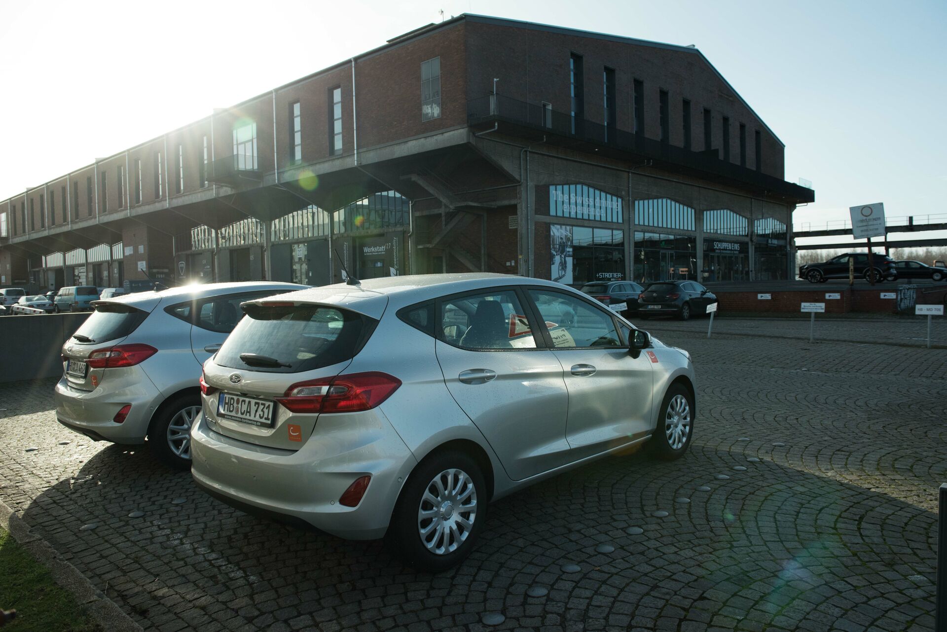 Im Vordergrund: Zwei silberne Autos. Im Hintergrund: Lagerhäuser