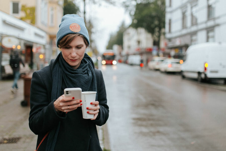 Frau steht an einer Straße, es ist regnerisch. Sie schaut auf ihr Handydisplay und hat in der anderen Hand einen Coffee-to-go-Becher.