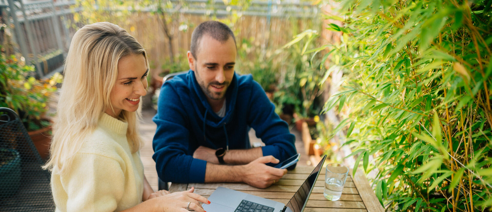 Eine Frau und ein Mann sitzen an einem Tisch auf einem Balkon und gucken fröhlich auf einen Computer