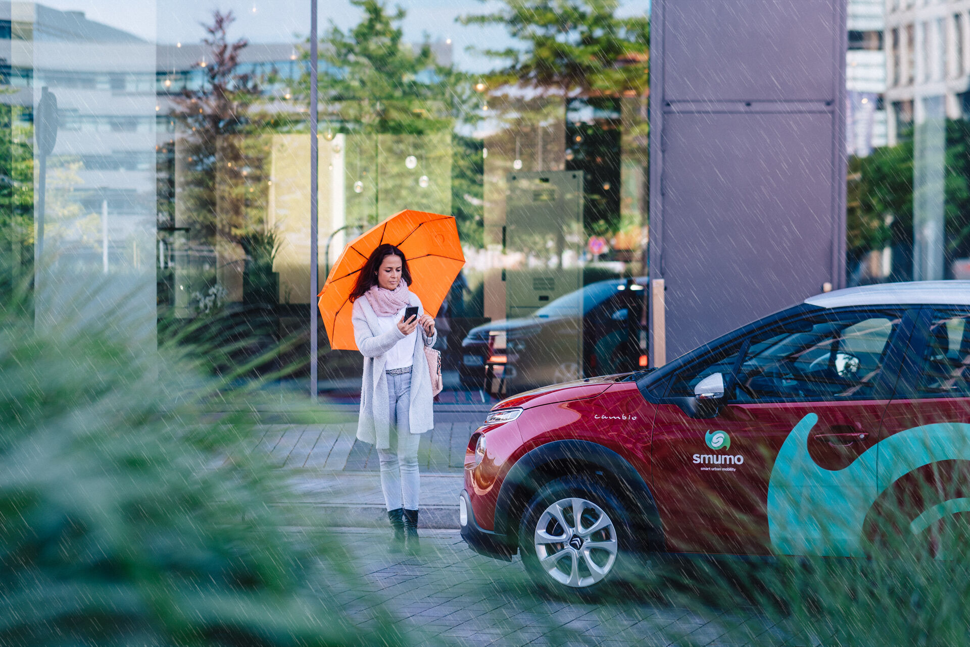 Frau mit orangem Regenschirm läuft an rotem Auto mir dem Schriftzug "smumo" vorbei.